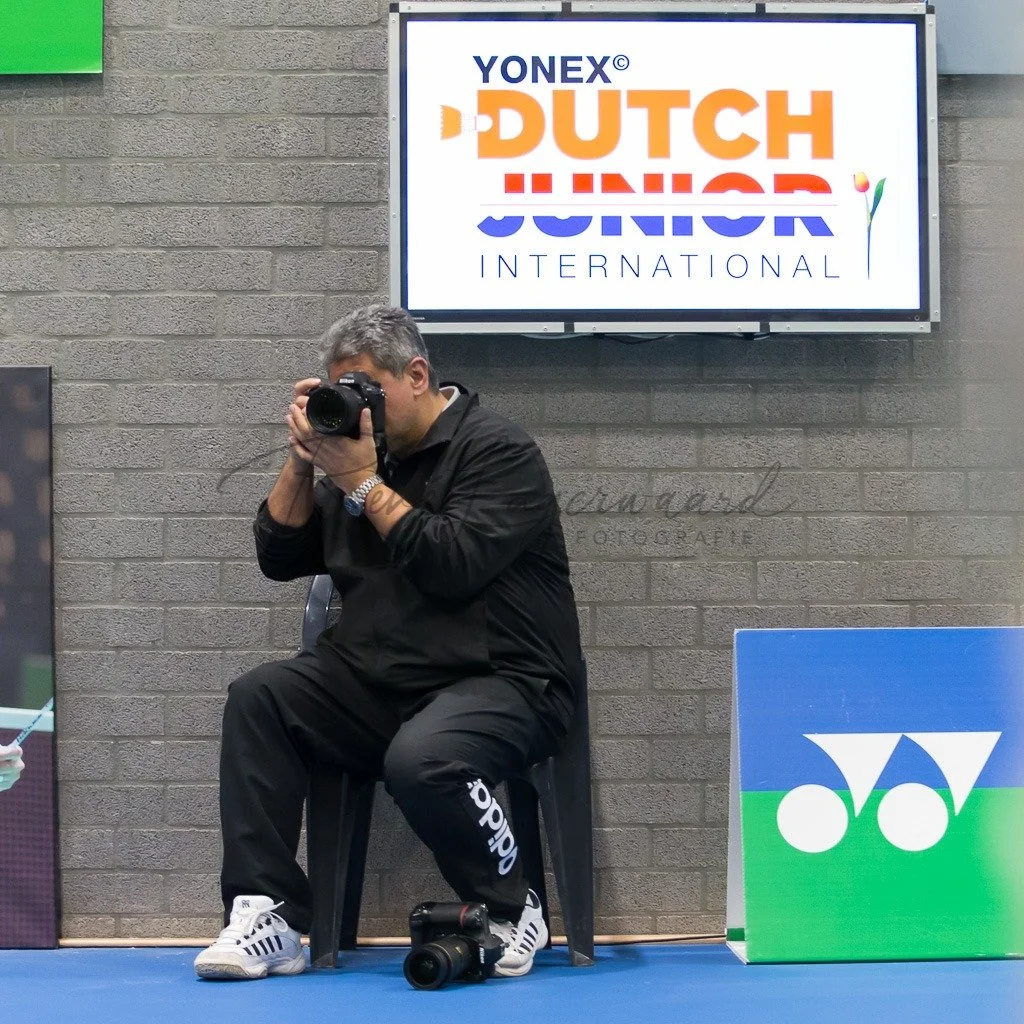 Man zit op een stoel en neemt een foto met een professionele camera. Op de achtergrond een tv-scherm met het logo van Dutch Junior International badminton.