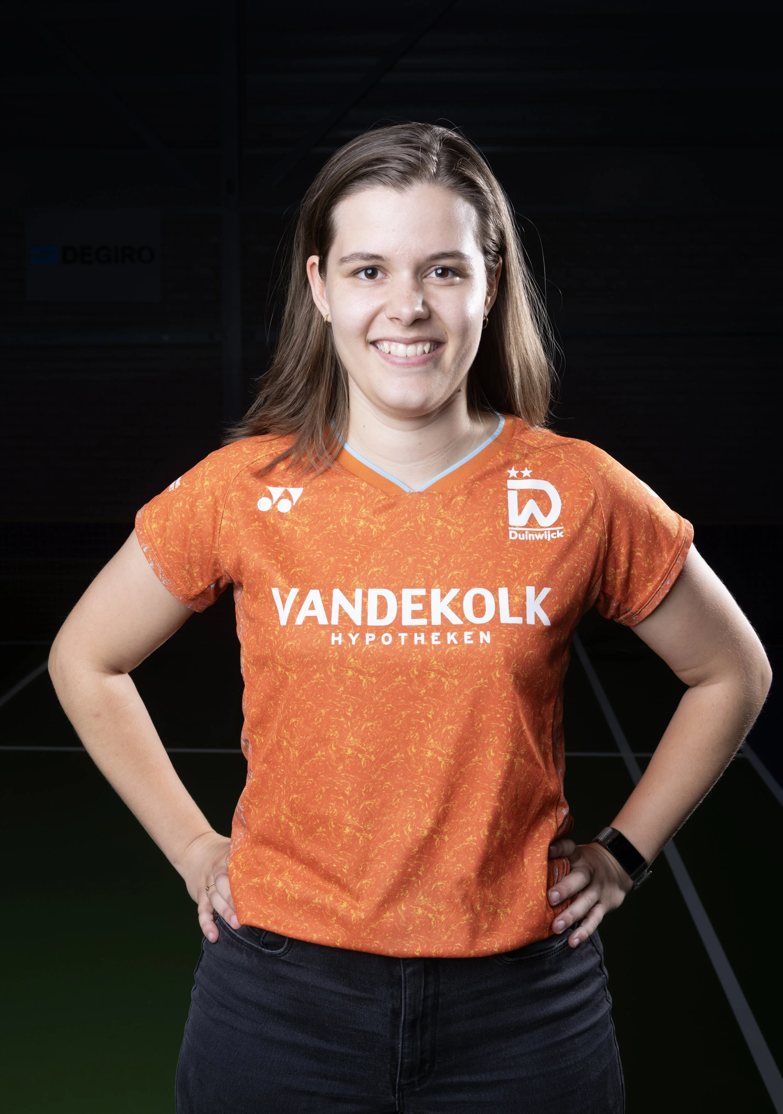 Laura de Quaasteniet, fysiotherapeut van BC Duinwijck, in oranje clubshirt met sponsorlogo's en smile, poseert met handen op haar heupen in de sporthal van BC Duinwijck.