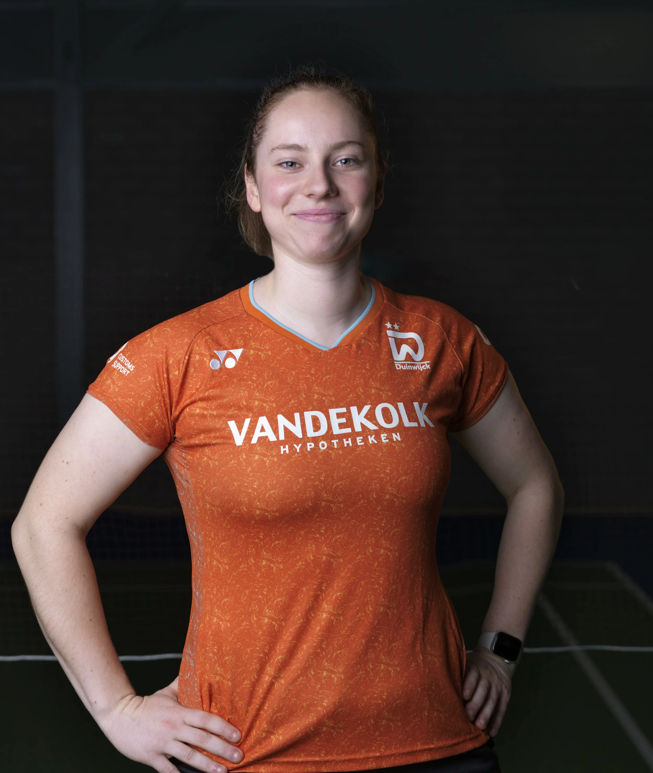 Close-up van een vrouwelijke atleet in oranje sportkleding met sponsorlogo's, poseert met handen op haar heupen in een sporthal.