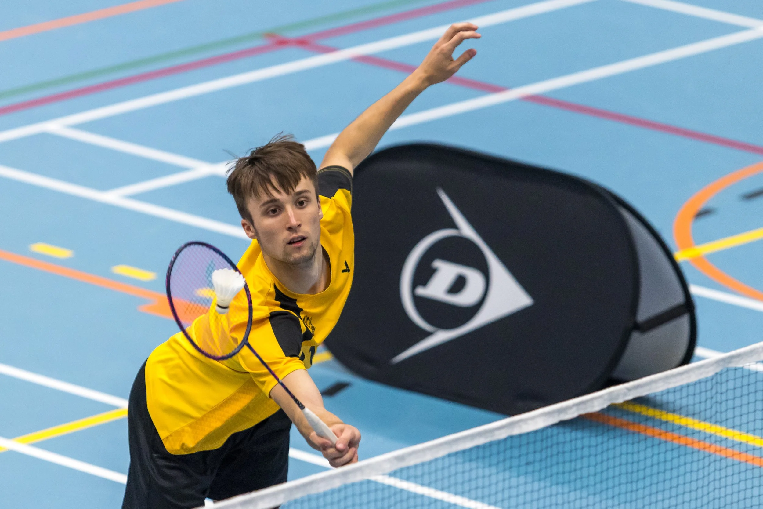 Dyon van Wijlick in een gele sportoutfit speelt badminton op een blauwe indoorbaan.