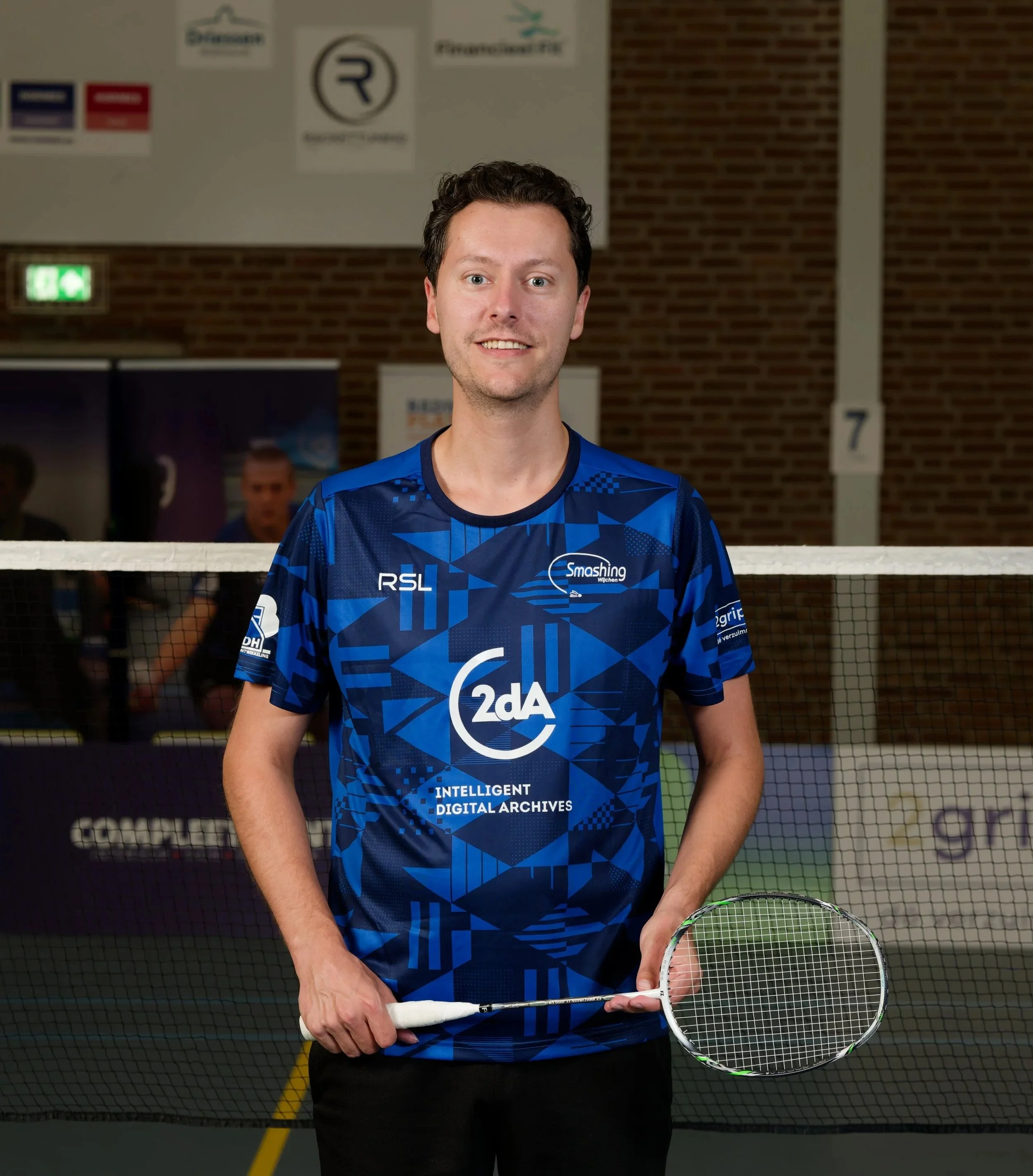 Man in een blauwe sportoutfit met badmintonracket in een indoor sporthal, achtergrond met net en posters op de muur.
