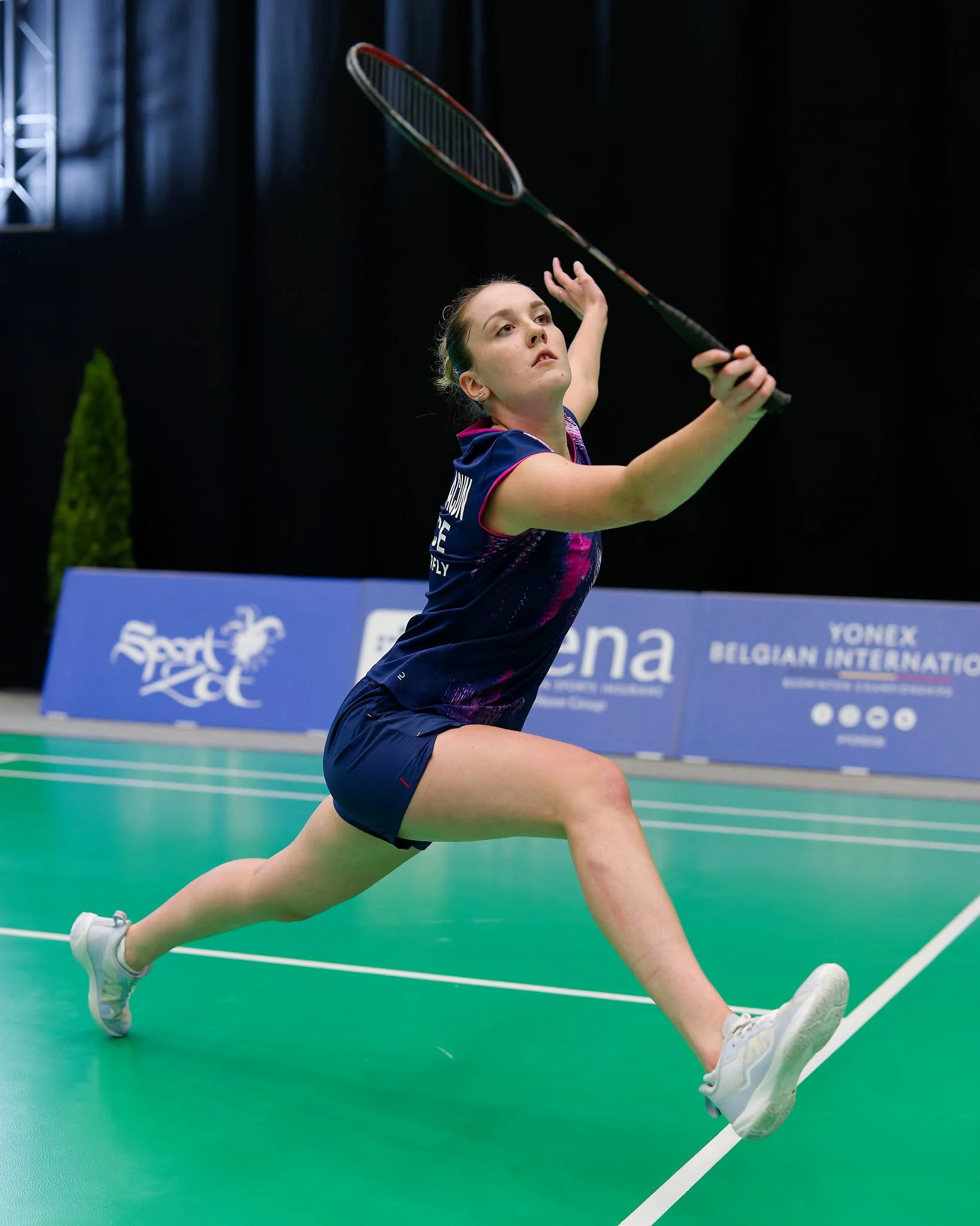 Kathell Desmots-Chacun, een vrouwelijke badmintonspeler in een blauwe sportoutfit in de beweging, die op haar knieën en met een badmintonracket voorbereidt om een volley te slaan op een indoor badmintonbaan.