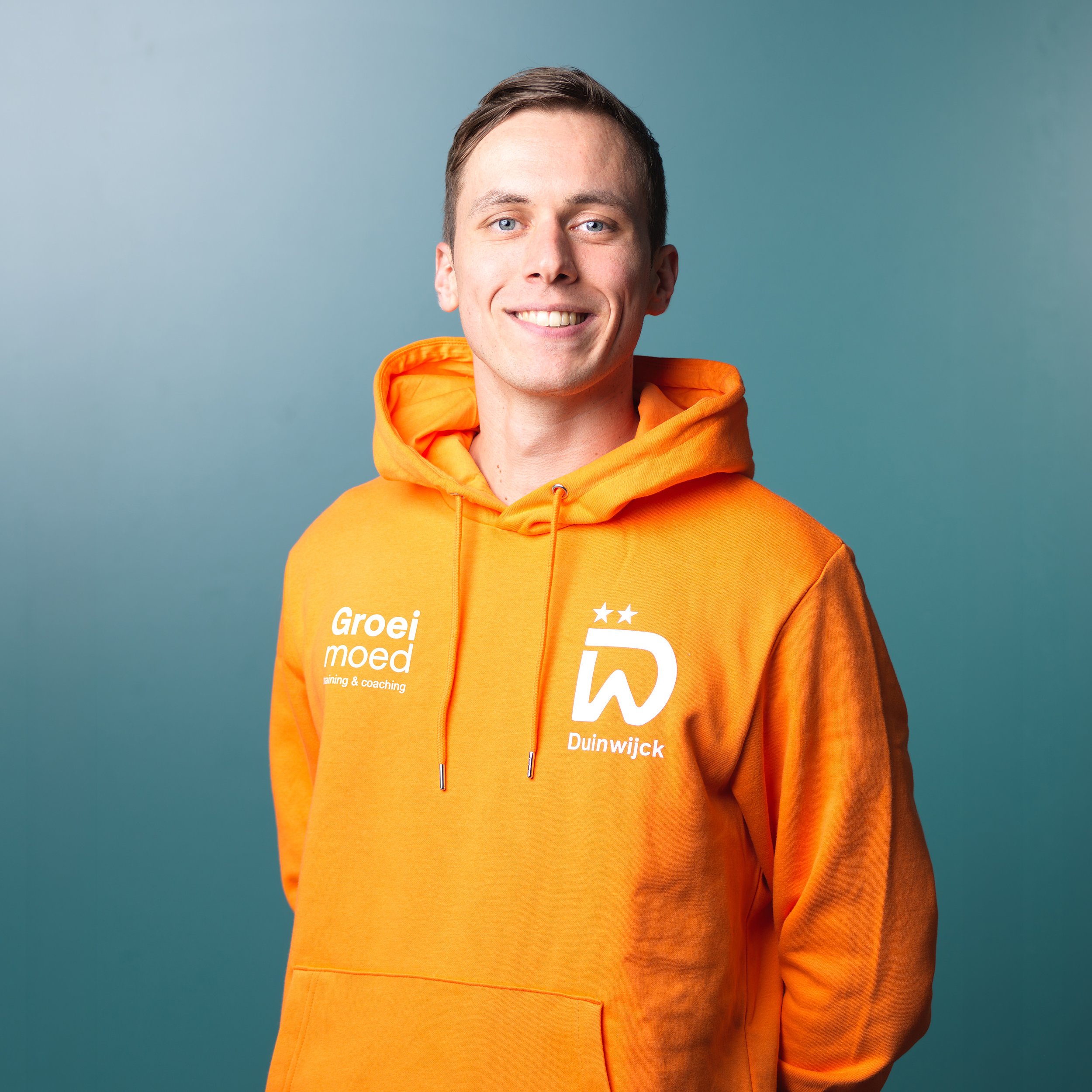 Close-up van een man in een oranje sportshirt met sponsorlogo's, armen over elkaar, glimlachend voor een donkere achtergrond.