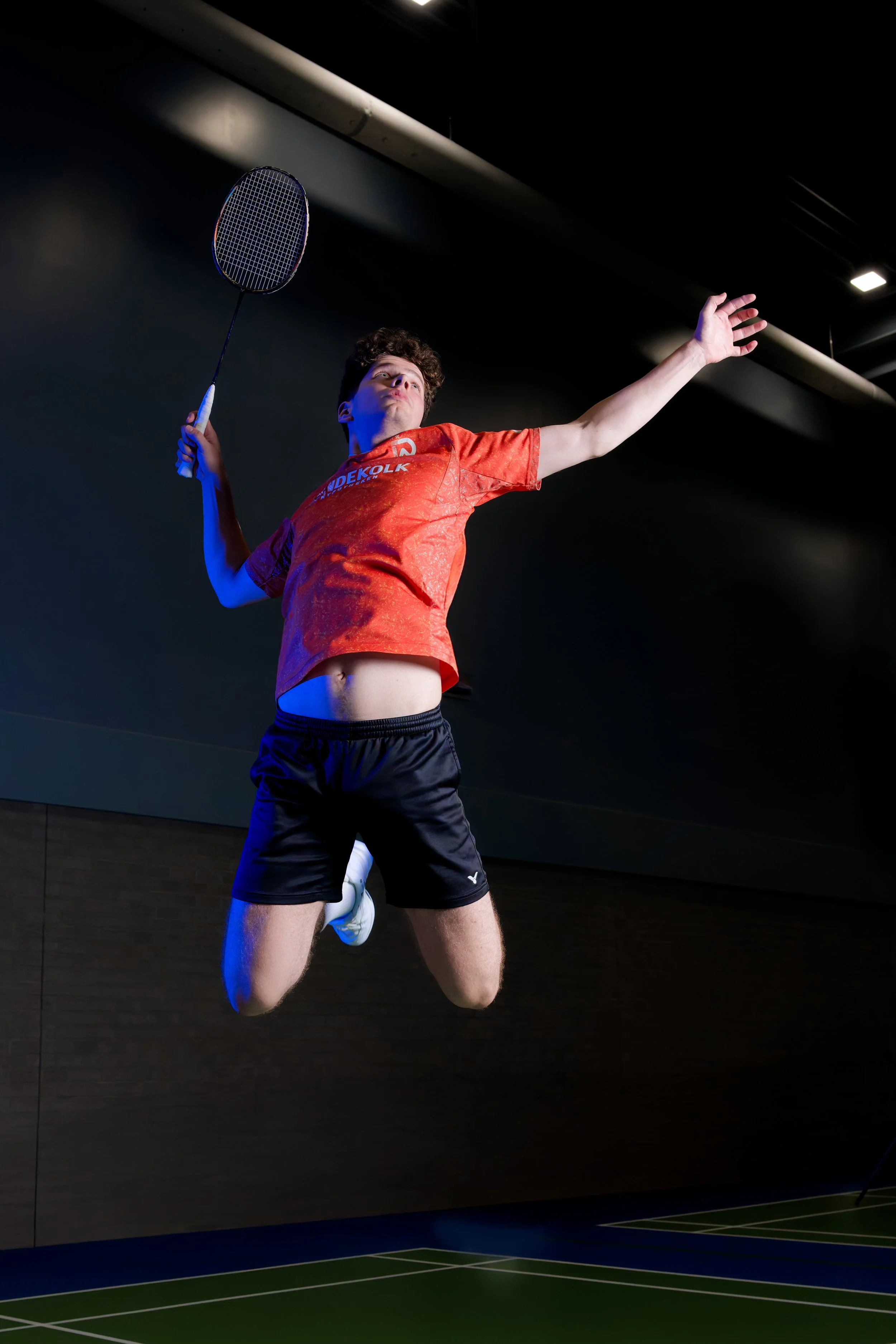 Een sporter in een sporthal, vastberaden in een sprong met een badmintonracket in zijn rechterhand, gekleed in een oranje sportshirt en zwarte sportshorts.