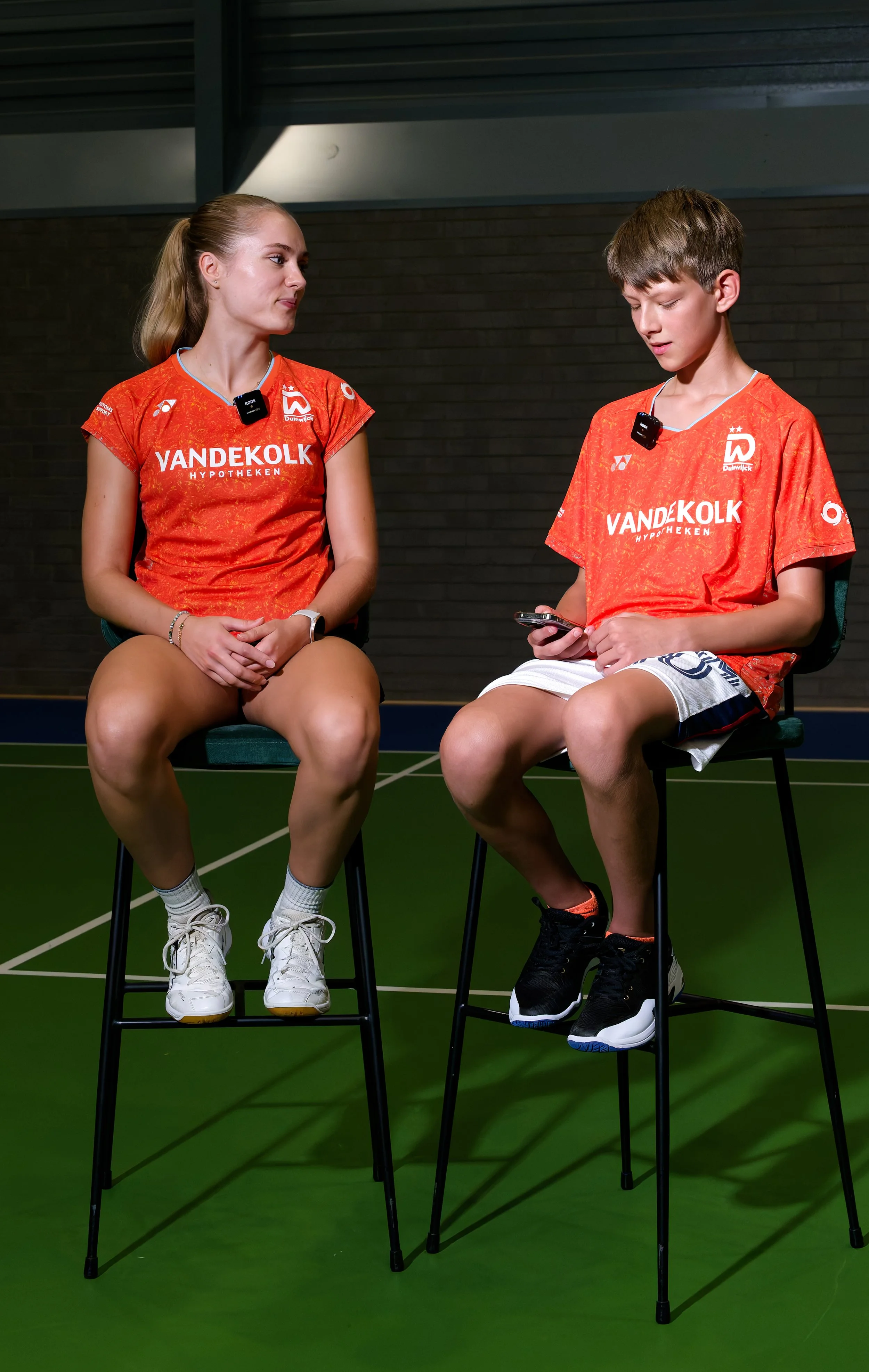 Twee jonge sporters in oranje shirts die in een sporthal zitten, de ene kijkt naar de andere die op haar telefoon kijkt.