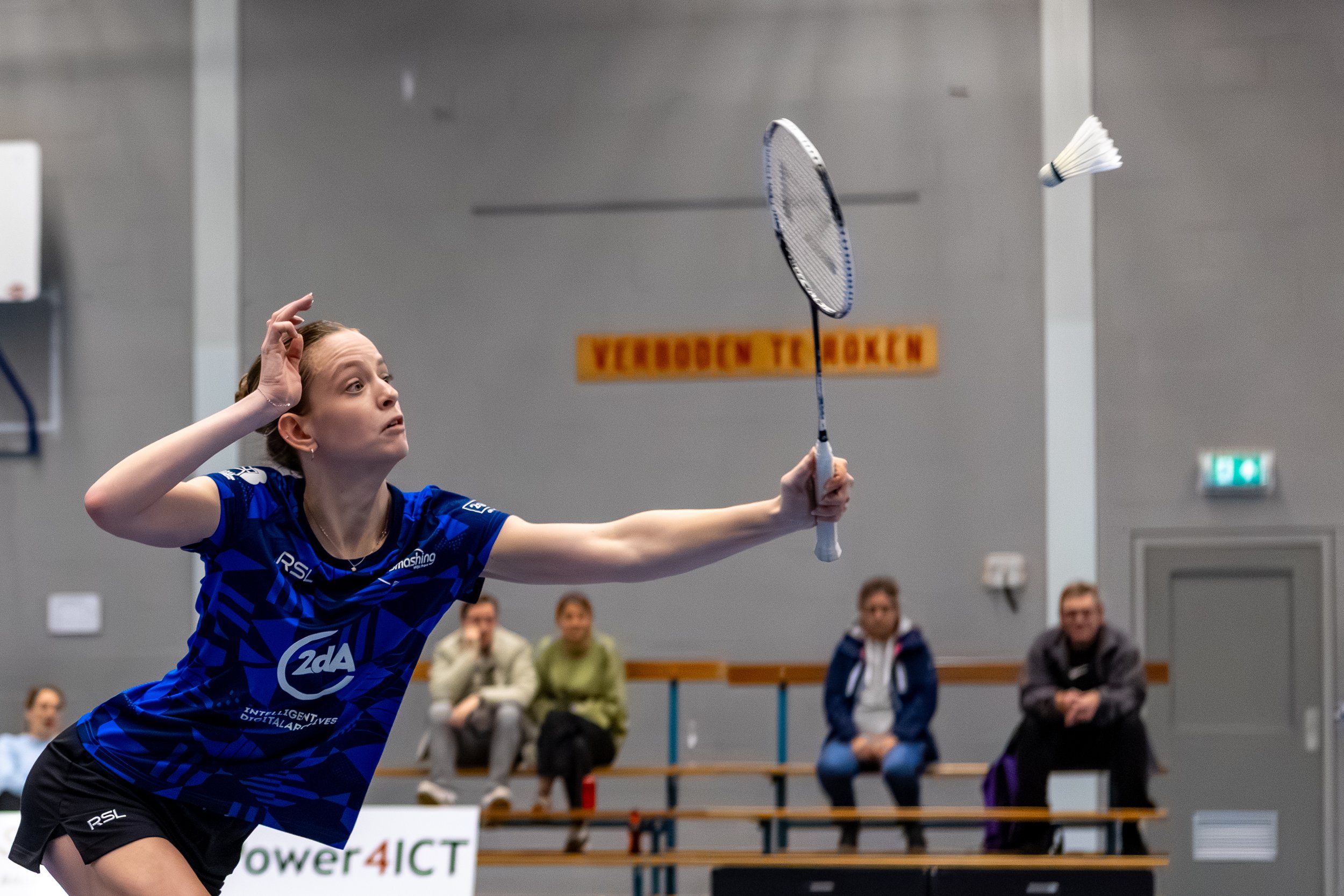 Sterre Bang speelt badminton in een sporthal, ze draait haar hoofd, houdt een badmintonracket in haar rechterhand en slaat een shuttle. Op de achtergrond zitten mensen op de tribune en kijkt men toe.