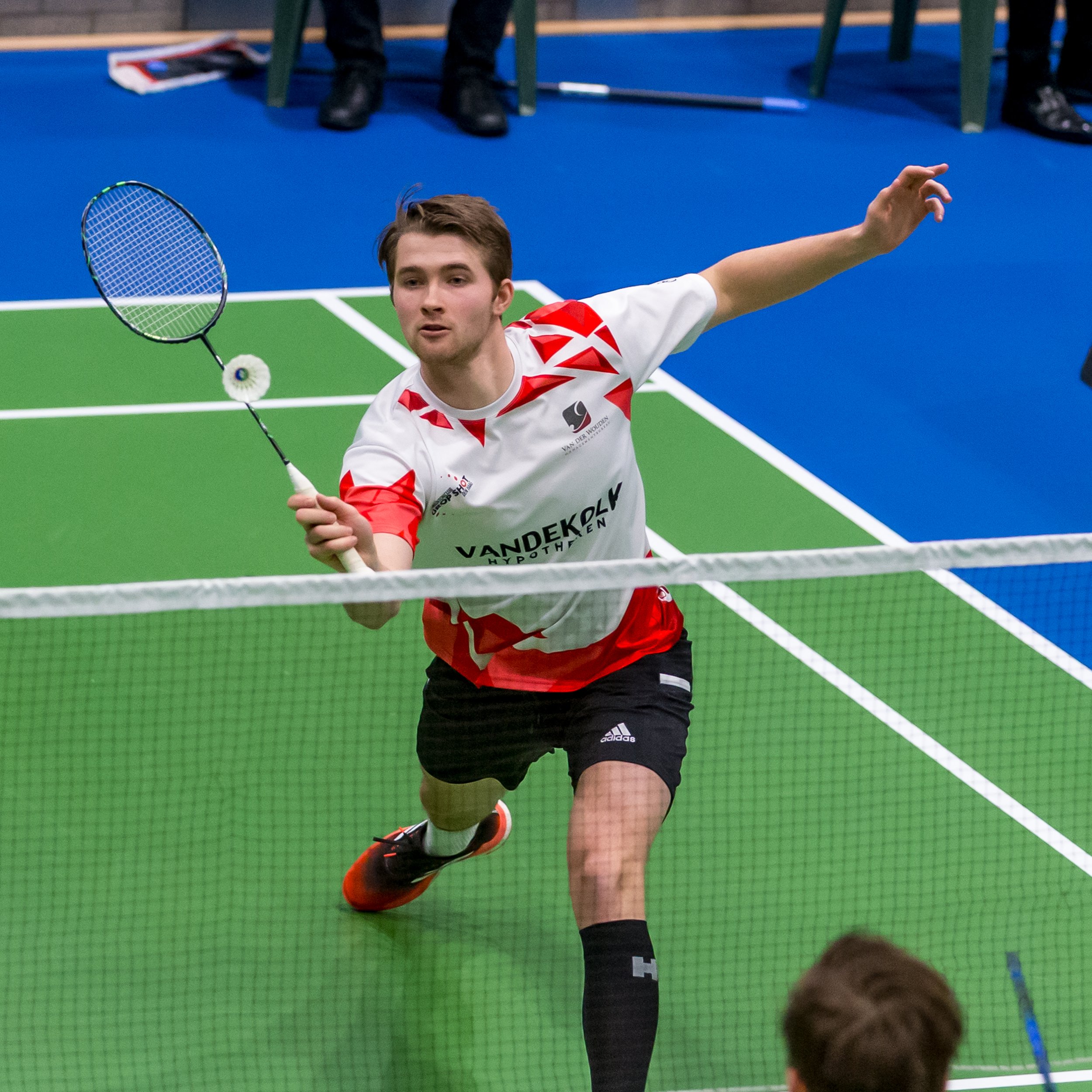Wessel van der Aar in actie tijdens een wedstrijd op een indoor baan. Wessel bevindt zich dicht bij het net en slaat een shuttle, terwijl de andere speler in de achtergrond klaar staat.