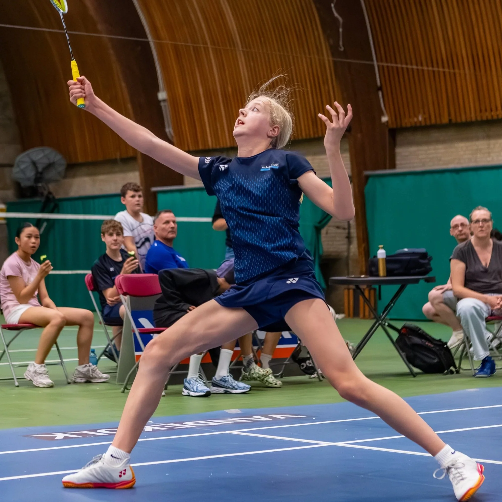 Meisje speelt badminton in een sporthal, met een groep mensen op de achtergrond.