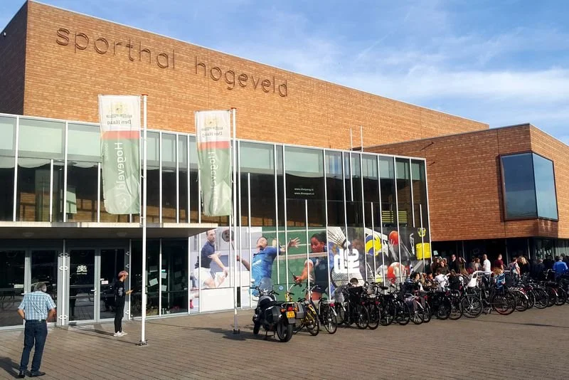 Het Sporthal Hoveveld, een sporthal met een moderne glazen gevel en fietsenstal buiten, met mensen die naar binnen en buiten gaan, onder een blauwe hemel.