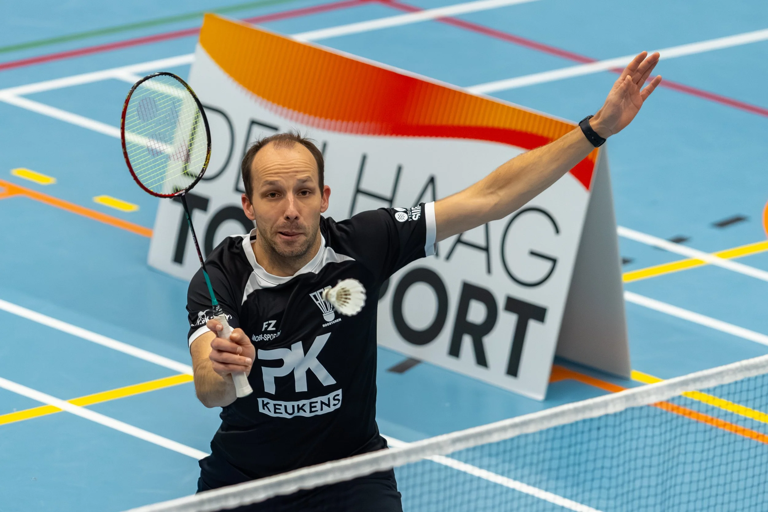 Alexander Roovers in zwart tenue slaat een shuttlecock tijdens een wedstrijd op een blauwe inside badmintonhal.