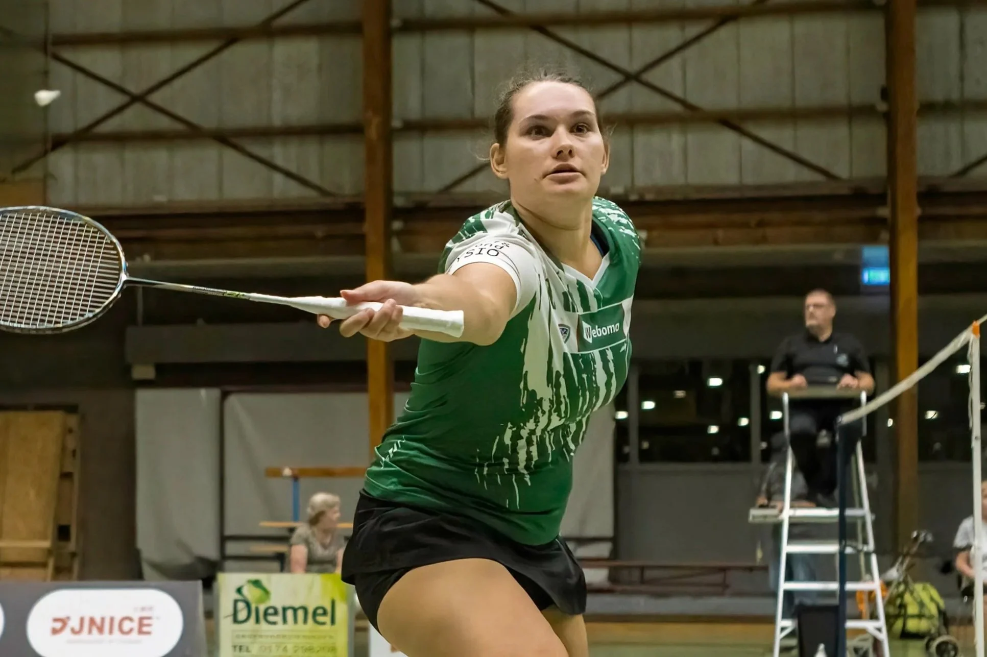 Myke Halkema, een vrouwelijke badmintonspeelster in een groene sportoutfit die naar de shuttle slaat in een binnenhal. Op de achtergrond zijn other mensen en wedstrijddoeken zichtbaar.