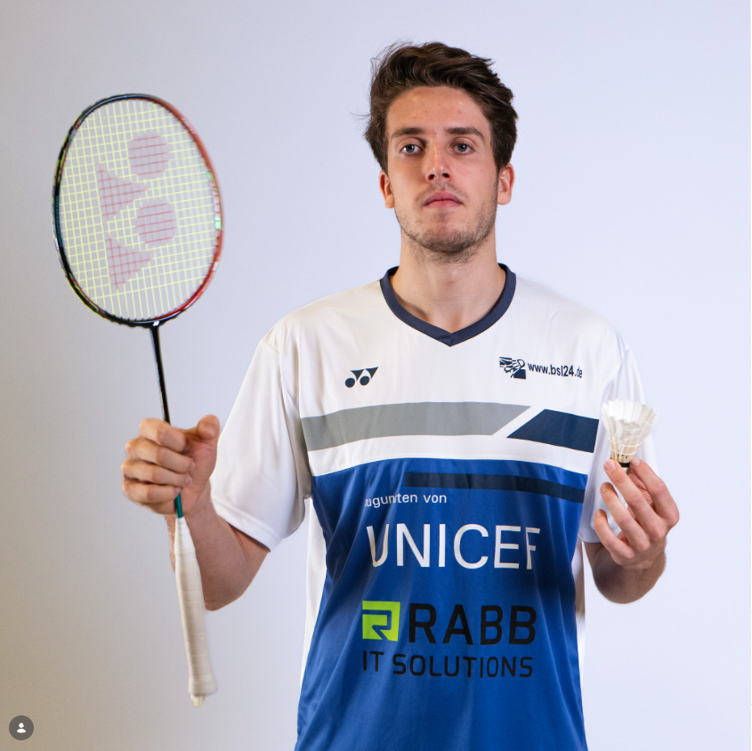 Marcello Kausemann in een badmintonshirt houdt een badmintonracket in de ene hand en een shuttle in de andere hand voor een witte achtergrond.