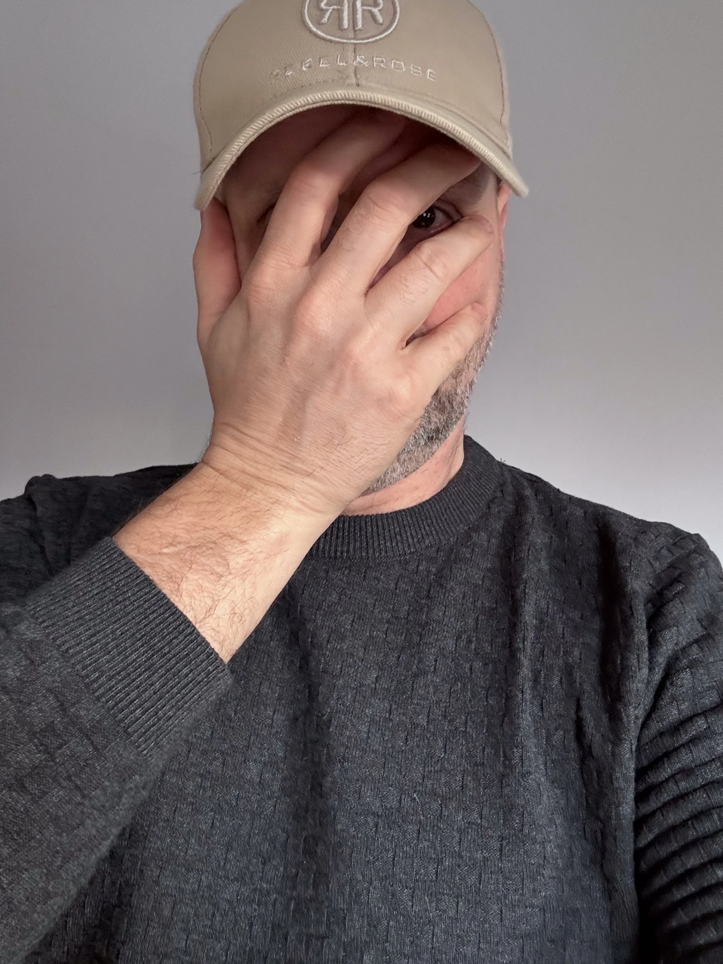 Man met een beige pet en een donkergrijze trui, die een hand voor het gezicht houdt, deels zijn gezicht bedekt en zichtbaar is slechts één oog.