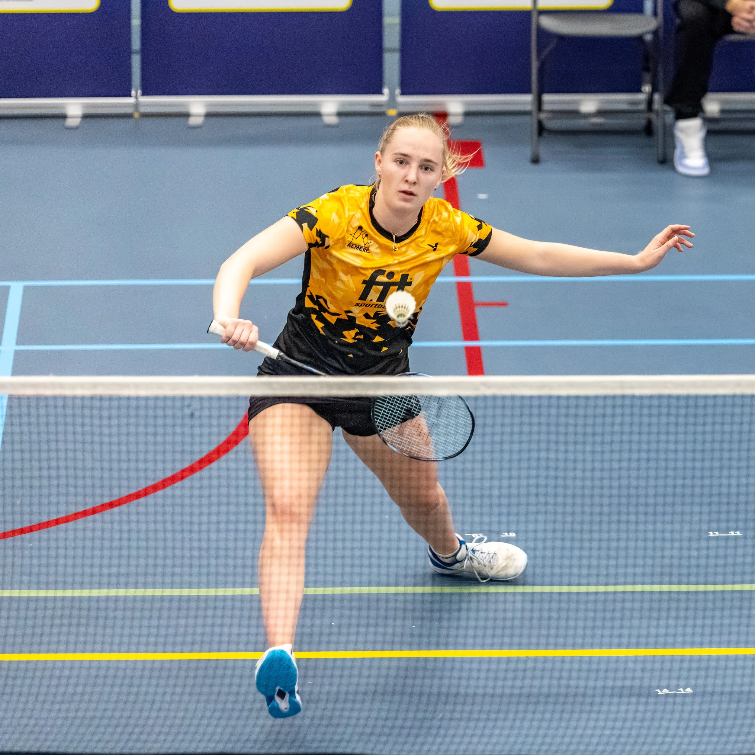 Kirsten de Wit op een indoor badmintonveld, met een badmintonracket in haar rechterhand en een shuttle in haar linkerhand. Ze kijkt geconcentreerd naar de shuttle terwijl ze zich voorbereidt op een slag.