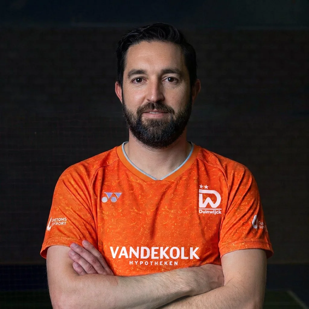 Man in oranje sportshirt met gekruiste armen, zittend voor een donkere achtergrond.