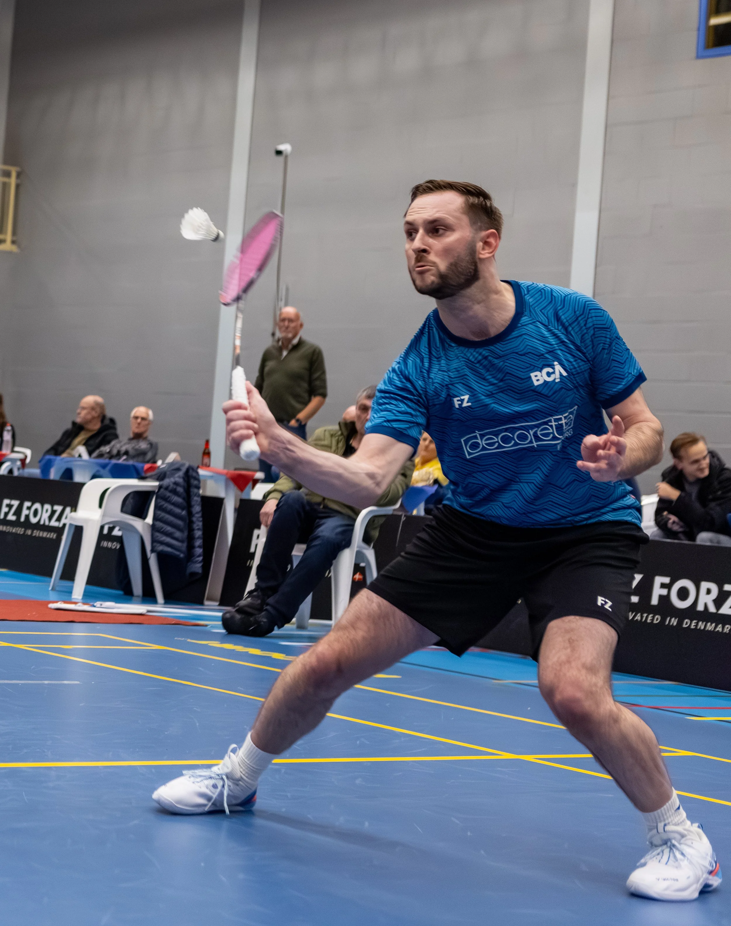 Ruben Jille in blauwe sportkleding speelt badminton in een binnenzaal, terwijl een andere man toekijkt.