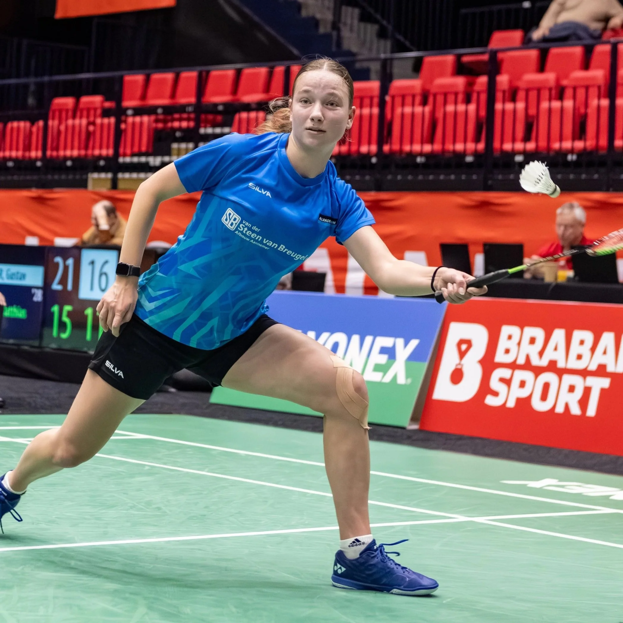 Mandy Duijndam in actie op een groene sportvloer in een indoor sporthal, gekleed in een blauwe sportoutfit en sportschoenen, met een badminton shuttle in haar hand.