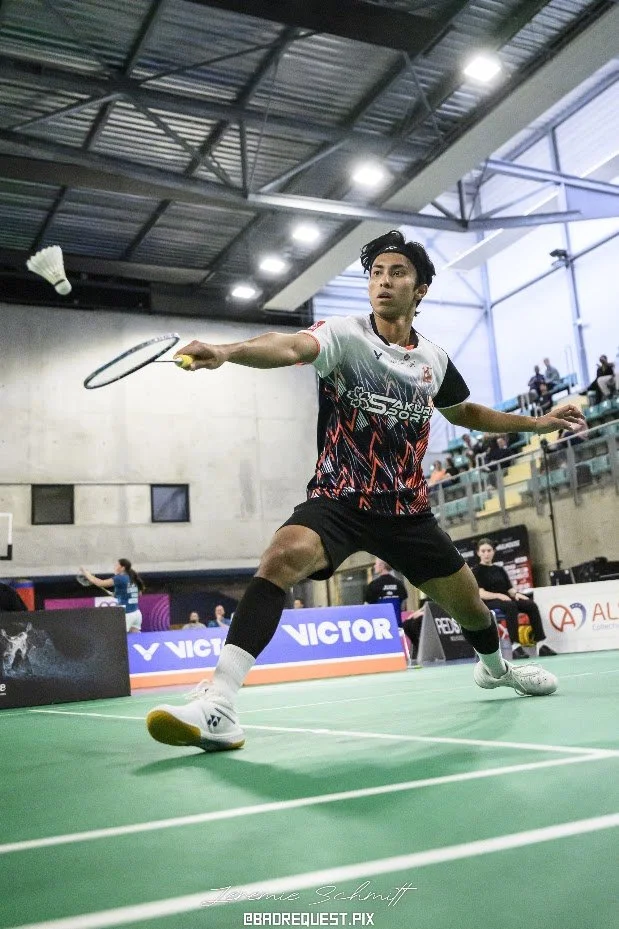 Een man speelt badminton in een indoor sporthal, hij is in een crouch positie die voorbereidt om de shuttle te slaan.