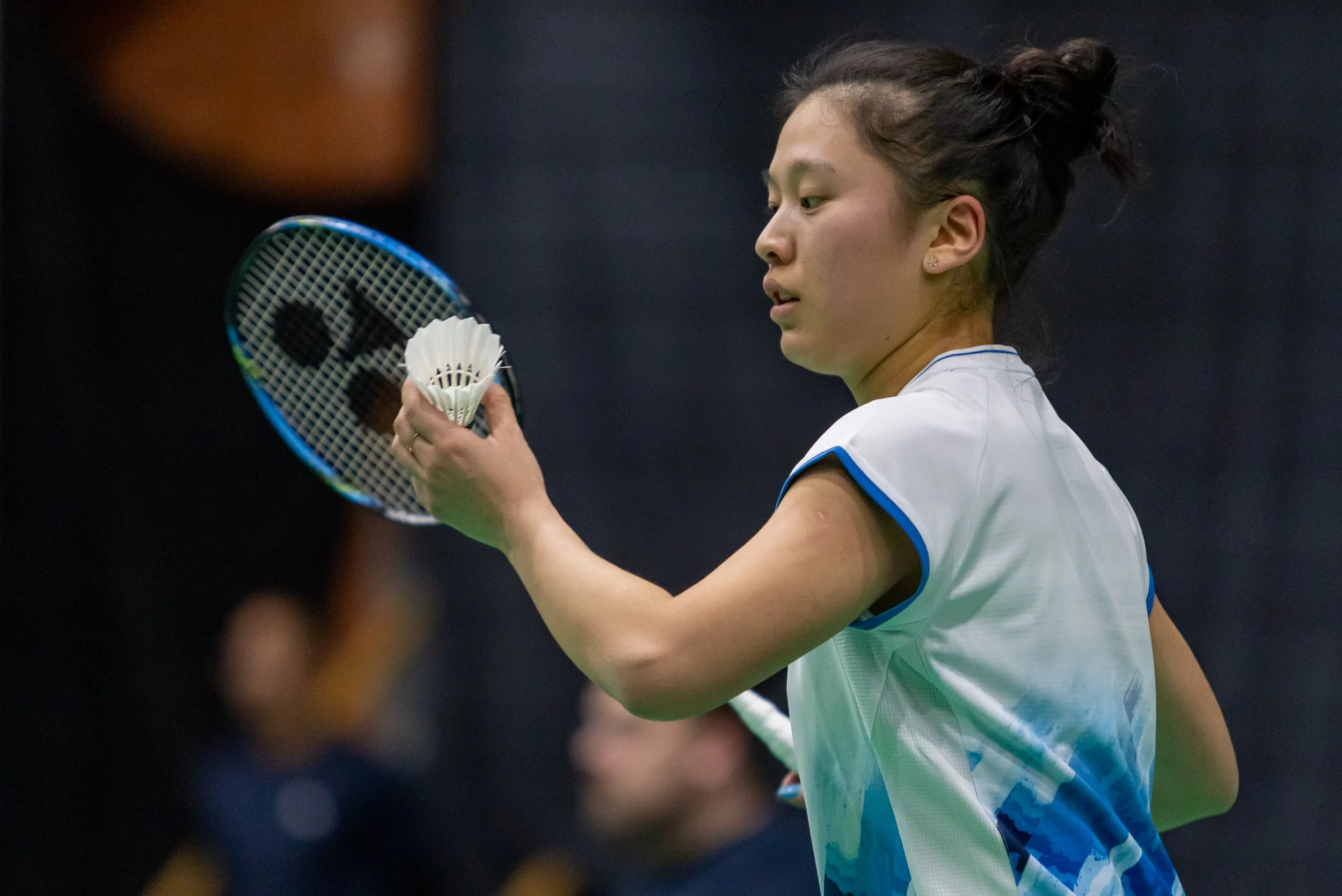 Amy Tan, een vrouwelijke badmintonspeler in een witte sportoutfit met blauwe details, klaar om te slaan, met haar haar in een knot, op een indoor badmintonbaan.
