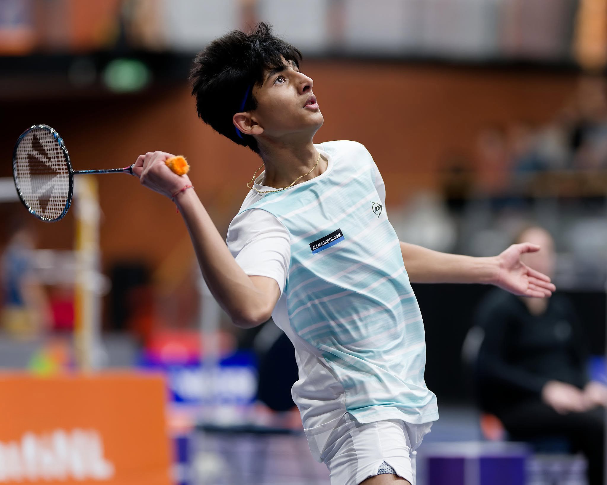 Vihaan Shah in een wit sportshirt met een badge en een goudketting speelt badminton in een sporthal, terwijl hij zich richt op het trekken van een slag.