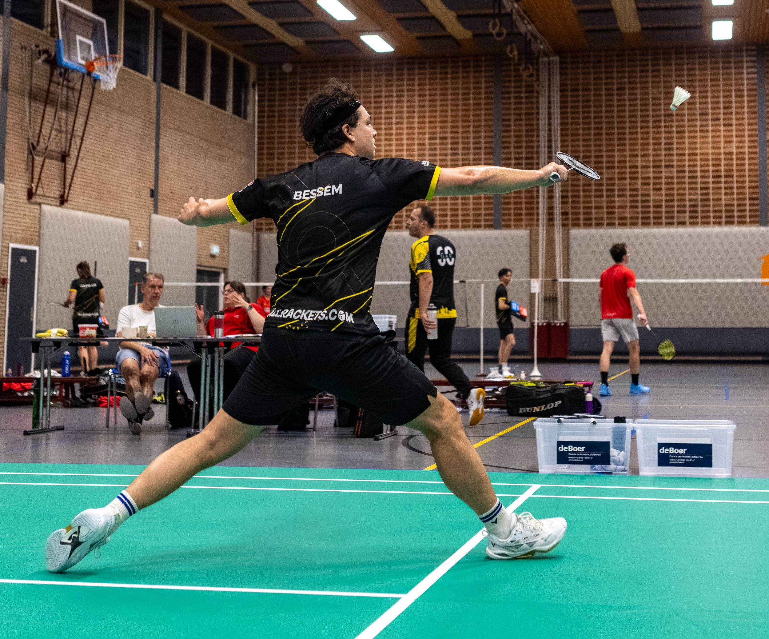 Brandom Bessem die badmintonspeelt in een sportschool, hij is in een uitvalpositie, met een rackets in zijn rechterhand en een shuttle die in de lucht zweeft.