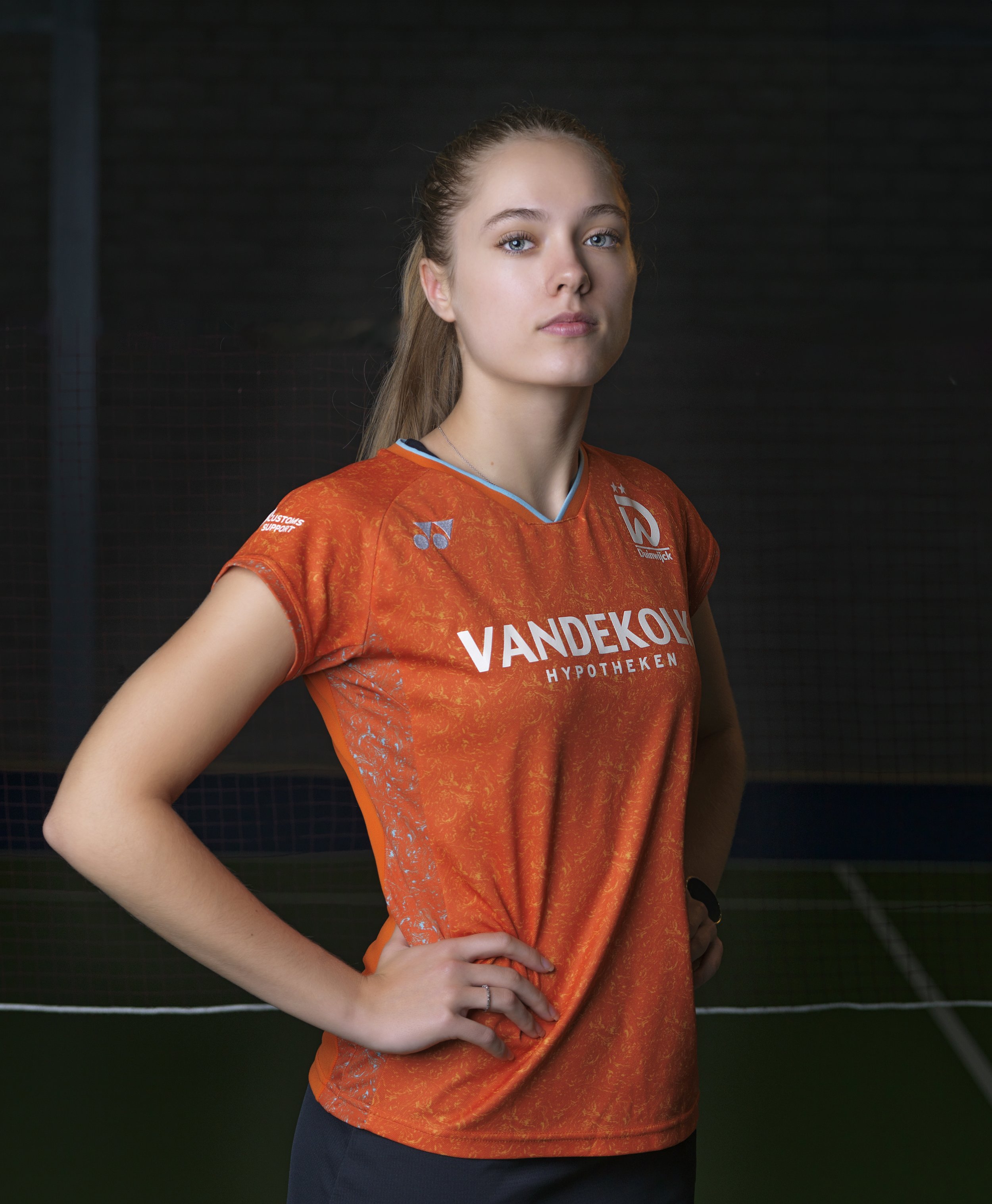 Sportvrouw in oranje sportshirt met sponsorlogo, poseert met hand op heup, binnen, donkere achtergrond.
