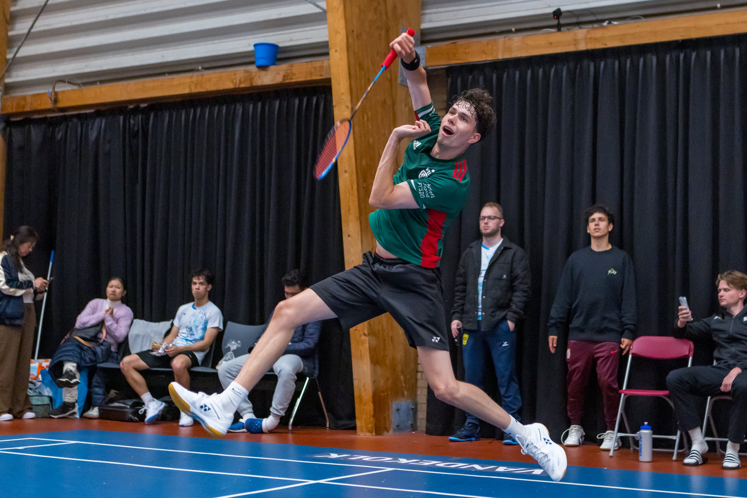 Mees Janssen speelt badminton indoors met een groene shirt en zwarte shorts, springend terwijl hij de shuttle slaat, op de achtergrond zitten mensen en kijken toe.