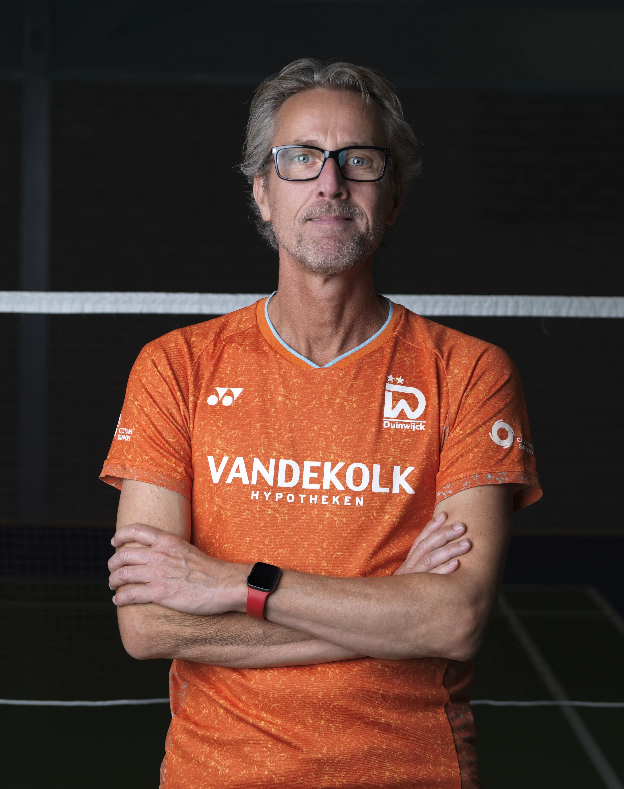 Man met grijs haar en bril, gekleed in een oranje sportshirt met sponsorlogo's, staat met gekruiste armen voor een donkere achtergrond.