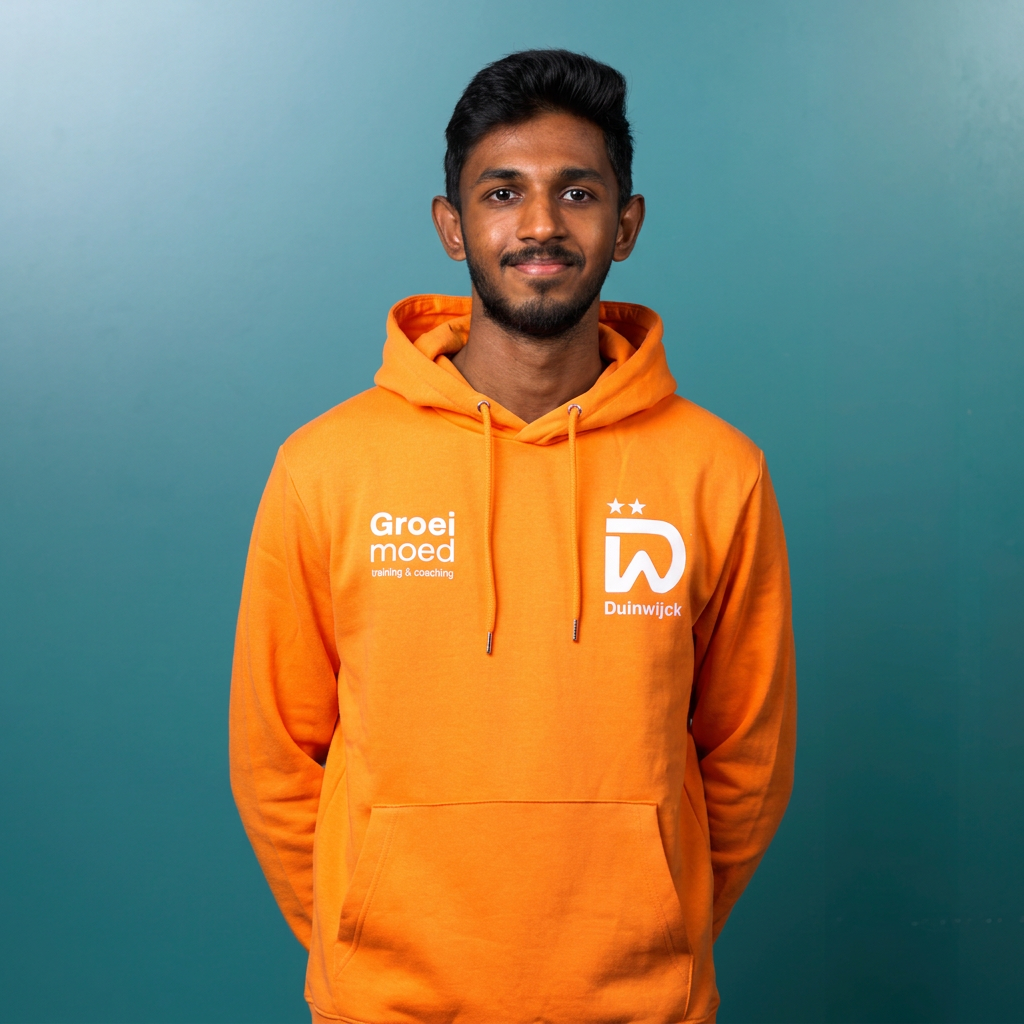 Close-up van een jonge man met een donkere huid en kort, golvend zwart haar, draagt een oranje sportshirt met witte en blauwe details, en staat met zijn armen over elkaar op een donkere achtergrond.