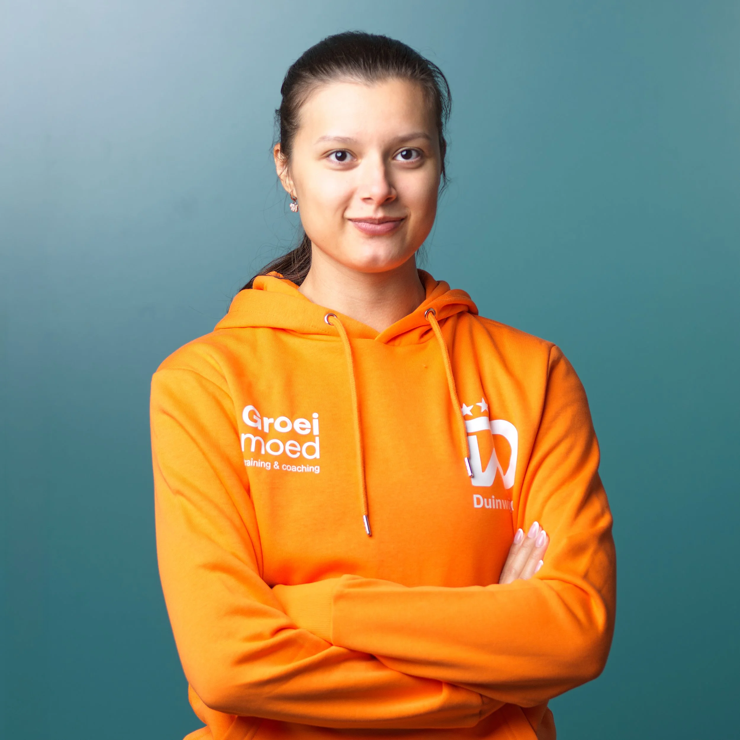 Close-up van een jonge vrouw met donker haar in een paardenstaart, oorringen, en een oranje sportshirt met tekst 'VANDEKOLK' en sportmerken, staand voor een zwarte achtergrond.