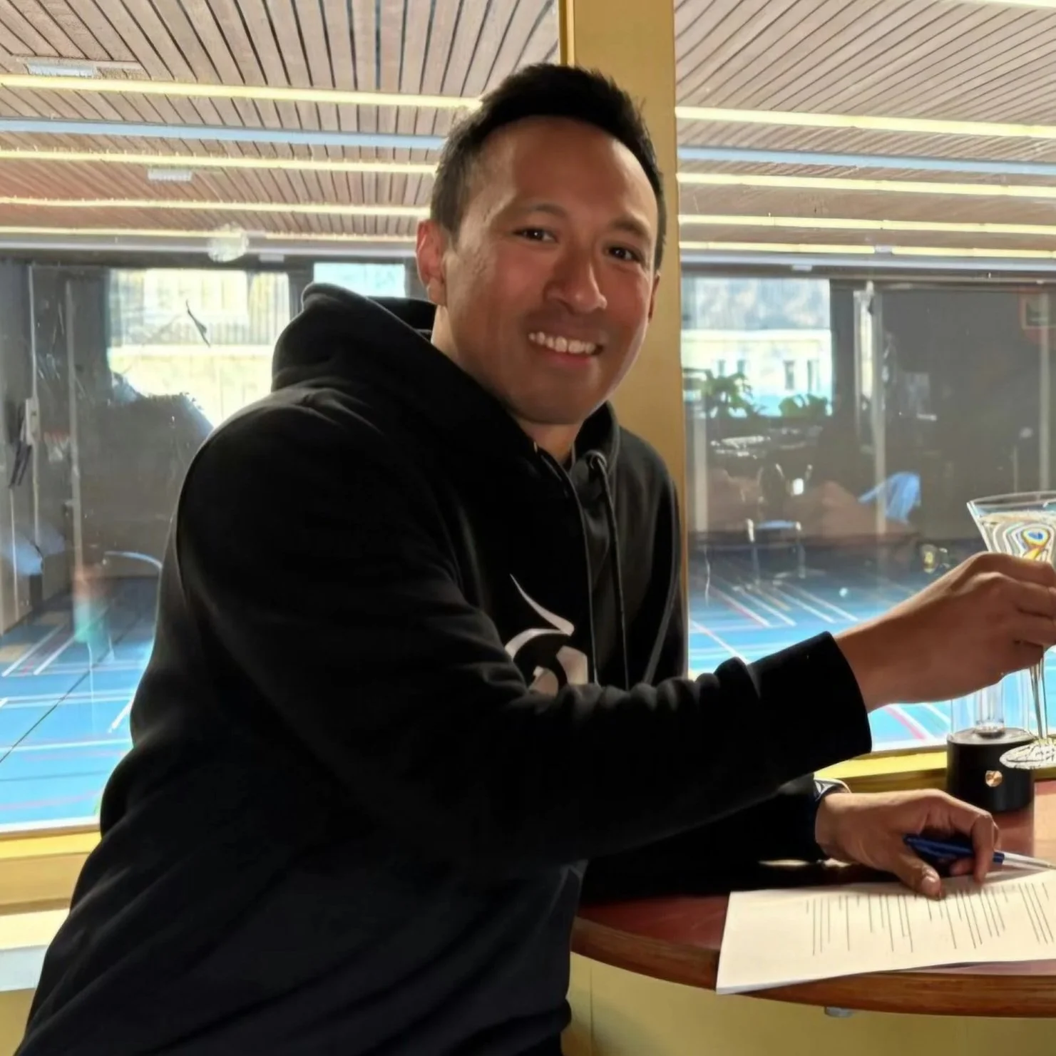 Man zit aan een tafel, houdt een glas in zijn rechterhand en heeft een document voor zich op de tafel, met een pen in zijn linkerhand. Achter hem grote ramen met een sporthal of gymzaal zichtbaar.
