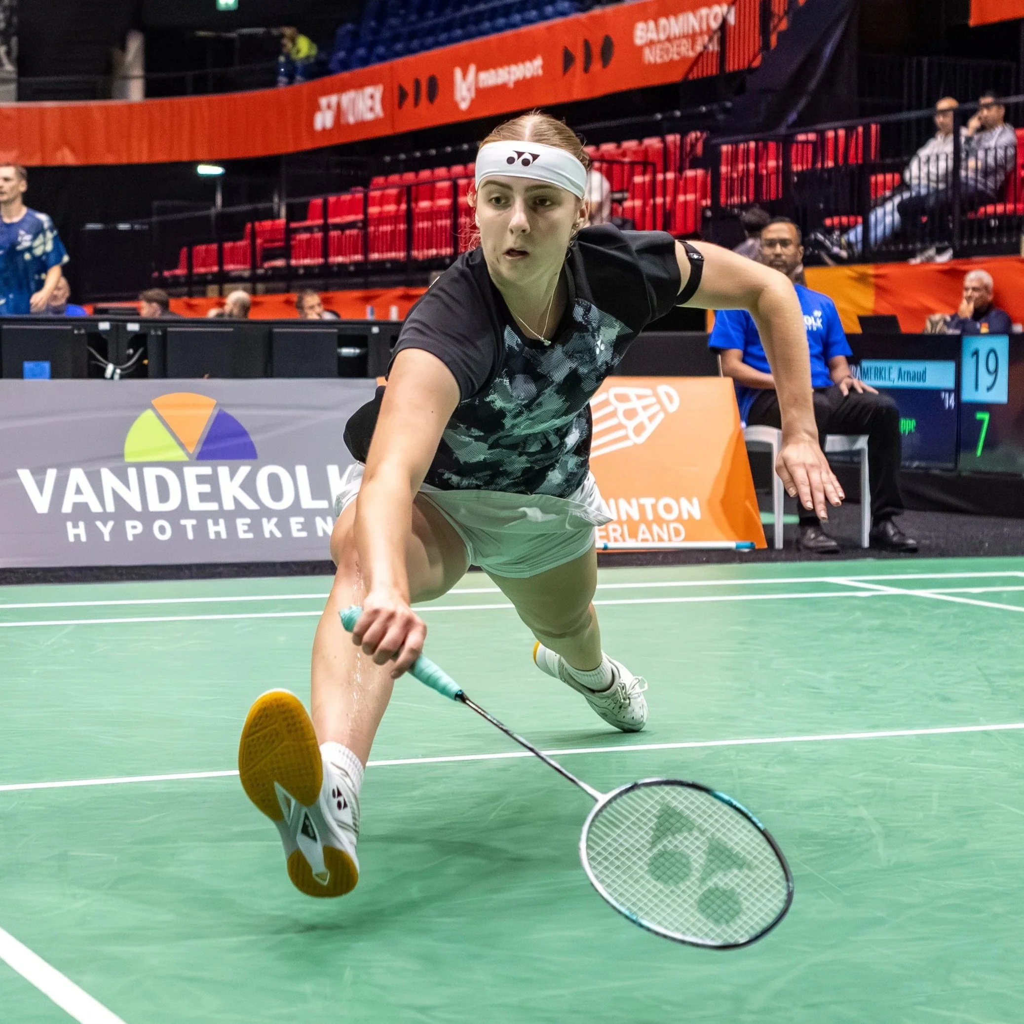 Tammi van Wonterghem in actie op een badmintonbaan, een kunstmatige vloer, met een gemiddelde publiek en banners op de achtergrond.