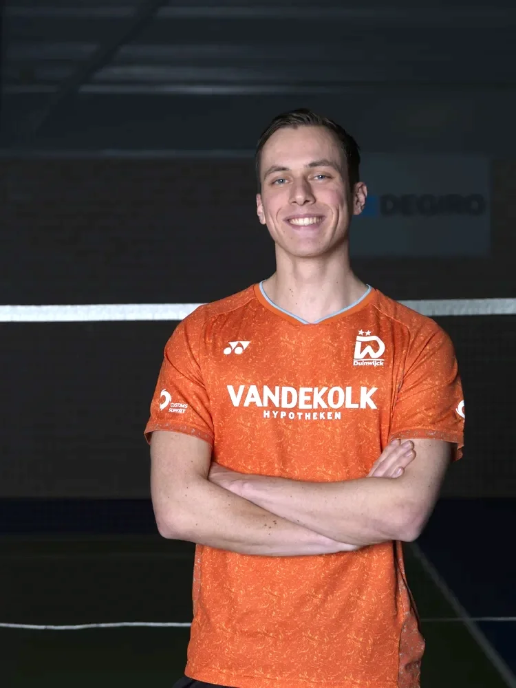 Brian Wassink in een oranje sportshirt staat binnen, lacht en heeft zijn armen over elkaar.