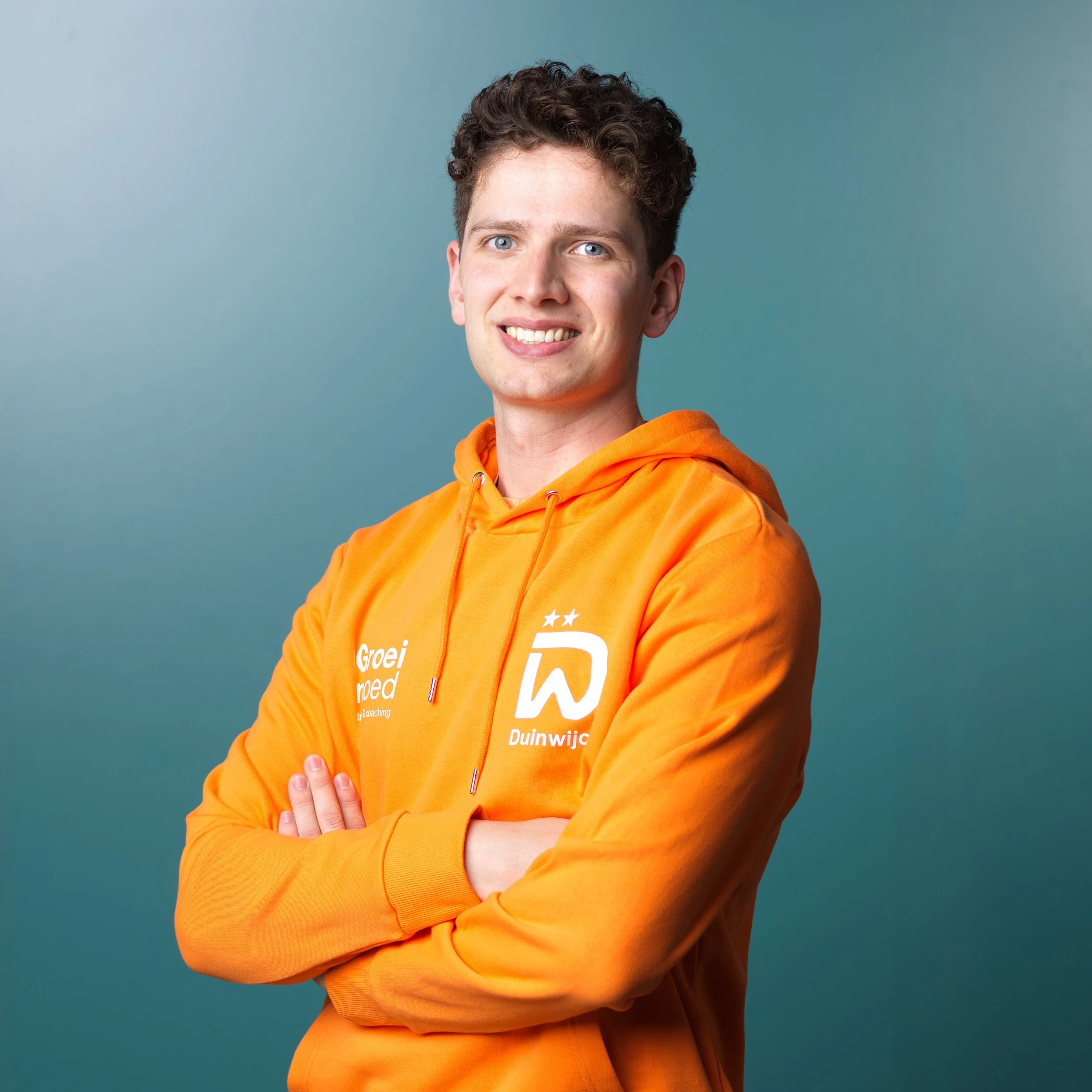 Een jonge man met blauwe ogen en kort, krullend haar draagt een oranje sportshirt met tekst en logo's, inclusief 'VANDEKOLK' en 'HYDROTHERAPY'.