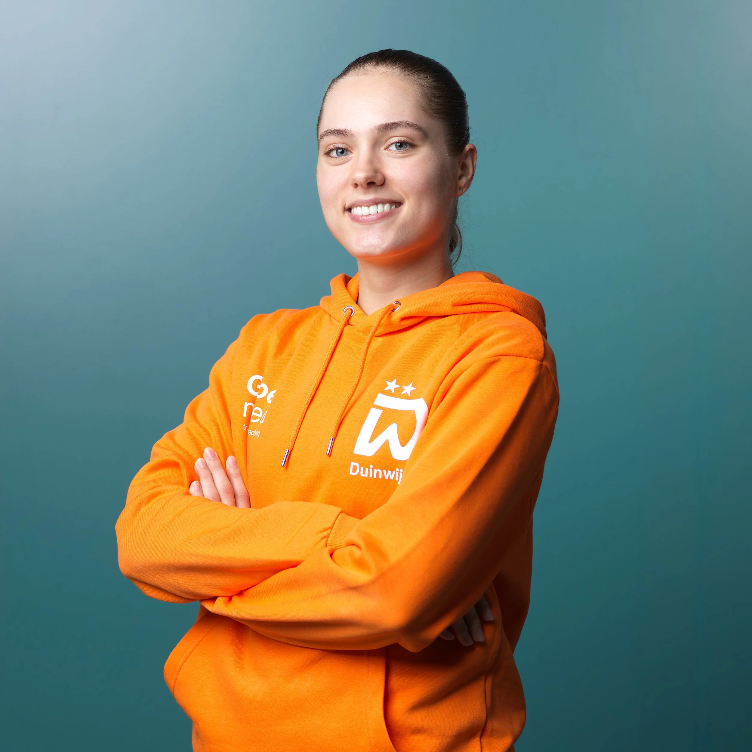 Een jonge vrouw met blond haar en blauwe ogen, gekleed in een oranje sportshirt met sponsorlogo's, staand voor een zwarte achtergrond.