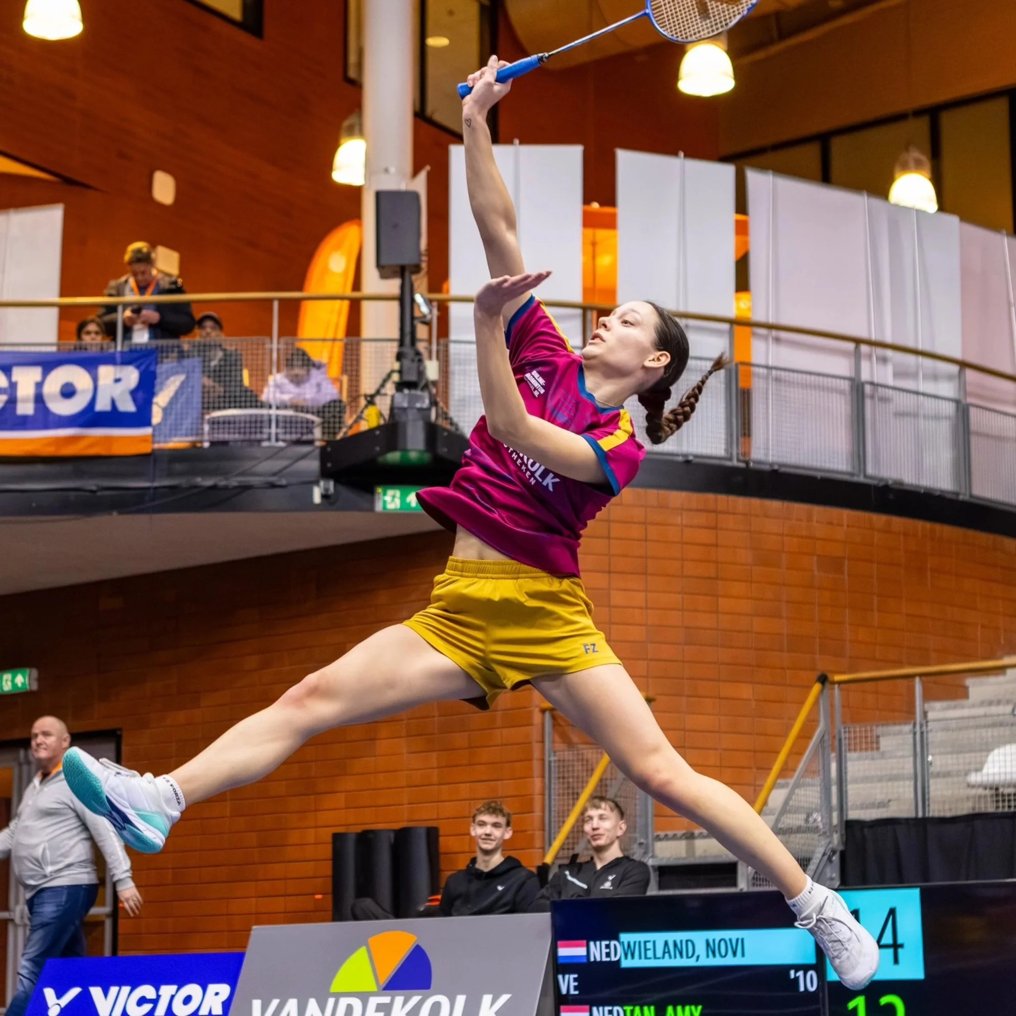 Novi Wieland in een roze en gele sportoutfit slaat de shuttle tijdens een match. Op de achtergrond kijken enkele mensen toe, waaronder drie jongeren die voor het scorebord zitten.