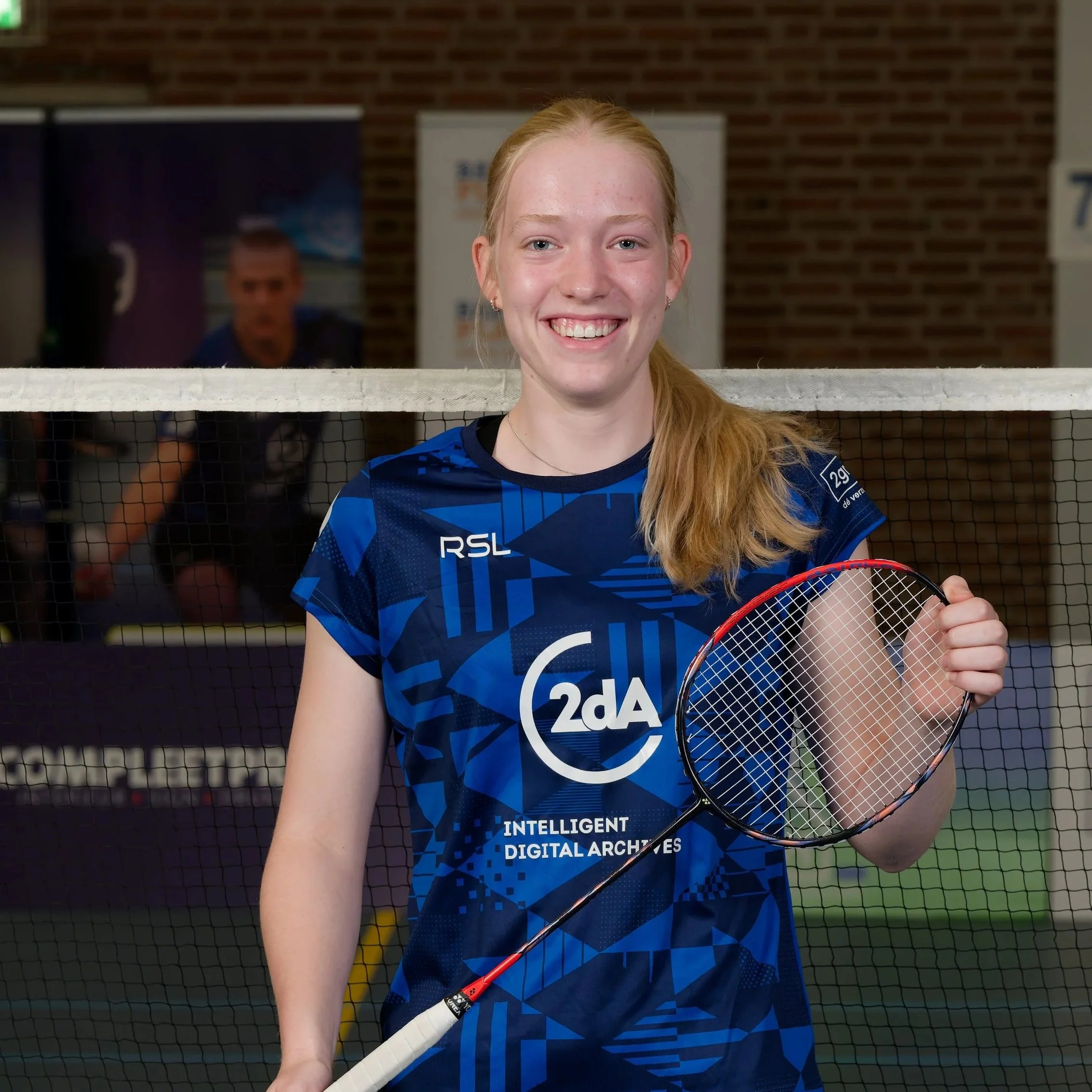 Een lachende jonge vrouw met rood haar in een blauw sportshirt met tekst 'INTEGRAAL SPORT' en een tennisracket vasthoudend, staat achter een tennisnet op een binnenbaan.