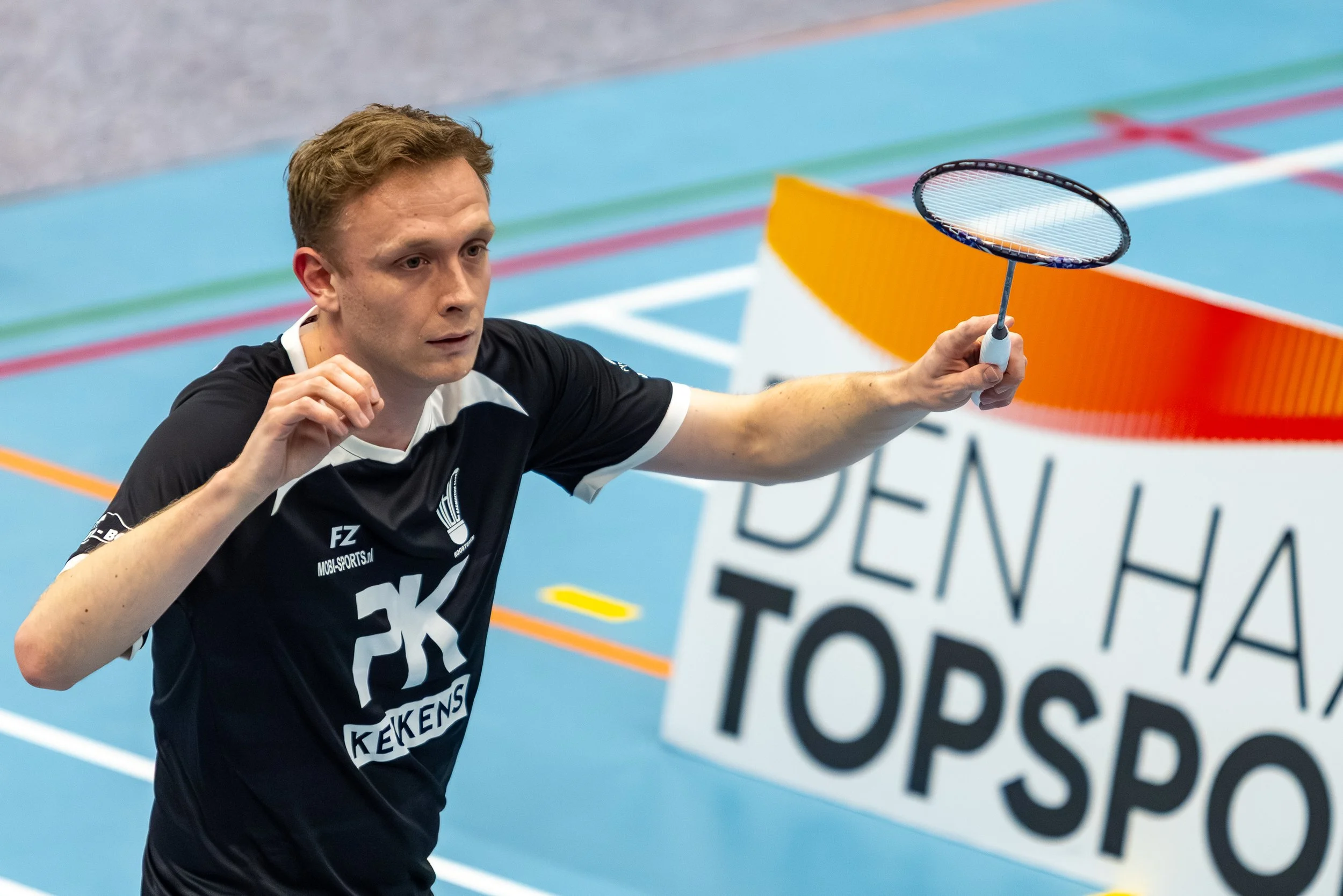 Jasper Koppen tijdens een badmintonwedstrijd, die zich richt en klaar is om te slaan met een racket in een sporthal met kleurrijke lijnen op de vloer.