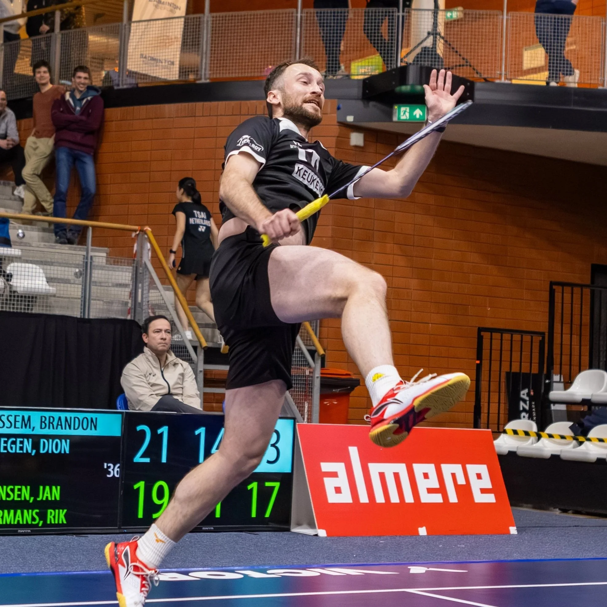 Een man in een zwart sportoutfit en witte schoenen met rode accenten springt over een hindernis tijdens een atletiekwedstrijd in een indoor sporthal. Op de achtergrond zien we een scoreboard en enkele toeschouwers.
