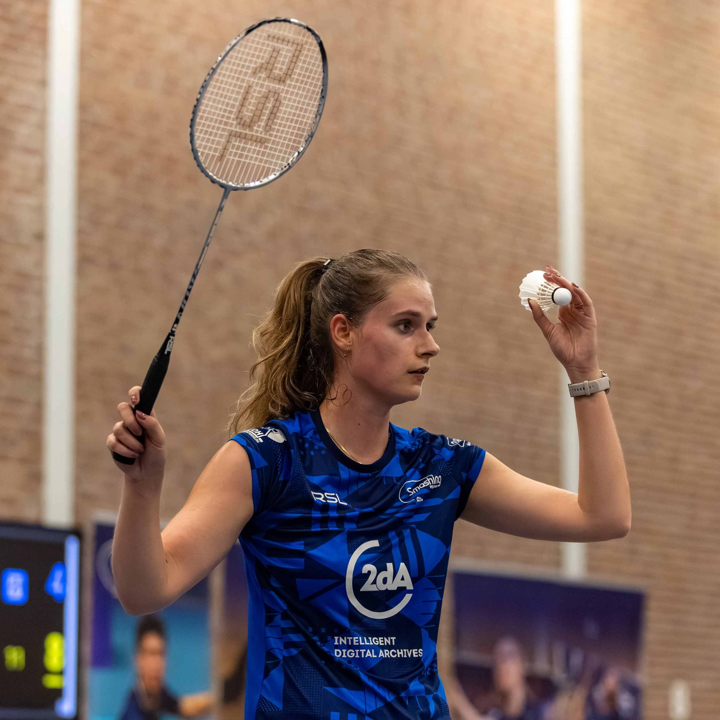 Maureen Zewald in een blauw sportoutfit die badminton speelt, bezig met het vasthouden van een shuttle en racket, binnen in een sporthal.