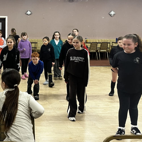 Group of children participating in an indoor activity or dance class in a room with tables and chairs along the wall.