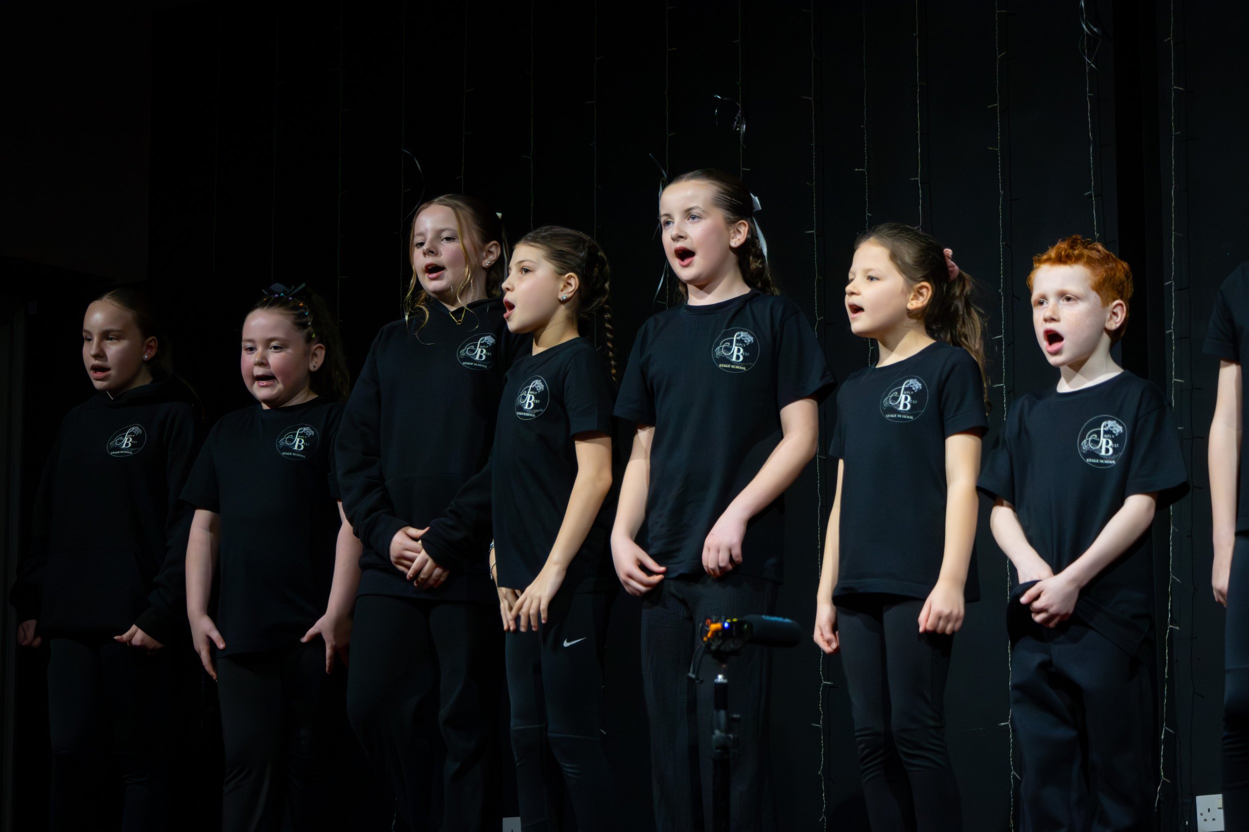 Group of young children singing on stage, wearing matching black shirts with a logo, with dark background and stage lights.