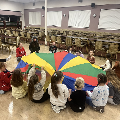 Children sitting in a circle playing with a colorful parachute in a large indoor room with stacked chairs and windows.