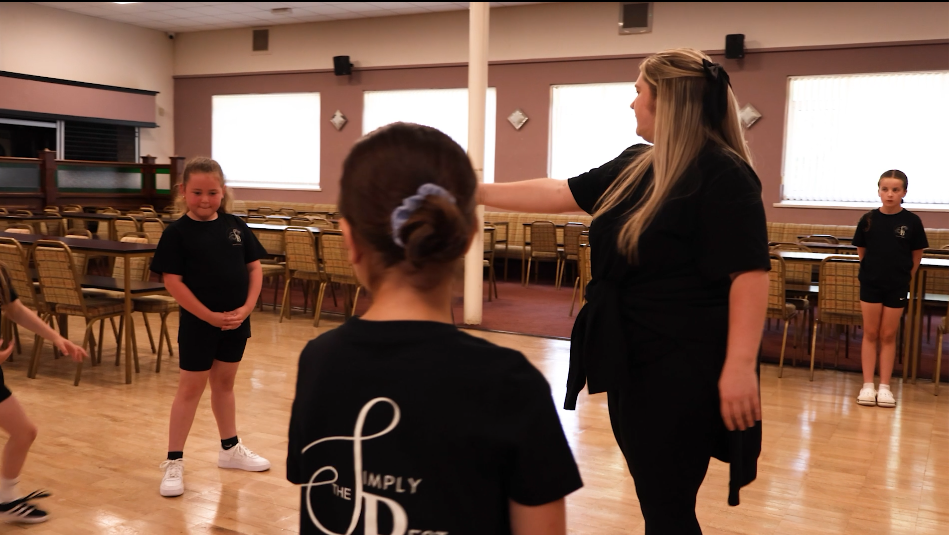 A woman instructing children in a dance or exercise class in a large room with wooden floors and empty chairs along the walls.