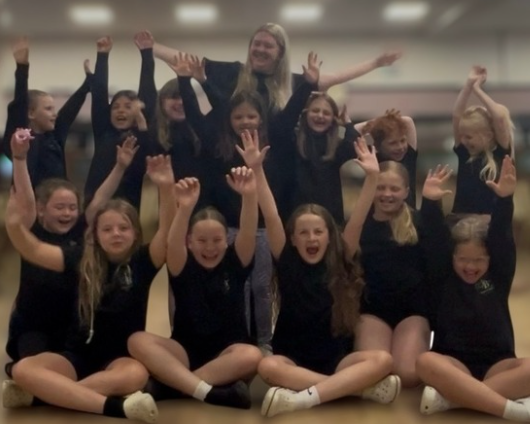 Group of young girls and a woman in a dance studio celebrating with raised hands and big smiles.