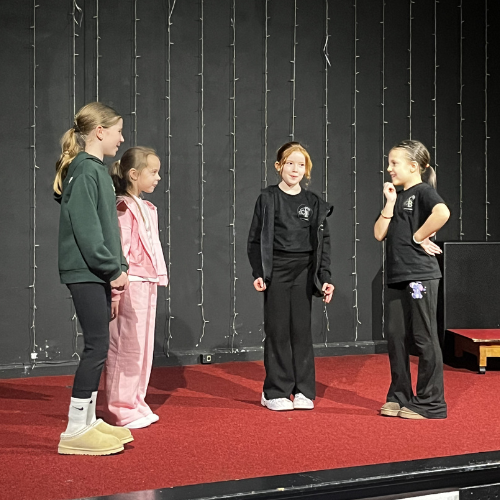 Four young girls standing on a red stage, engaged in a theatrical or school performance, with a black wall and string lights in the background.