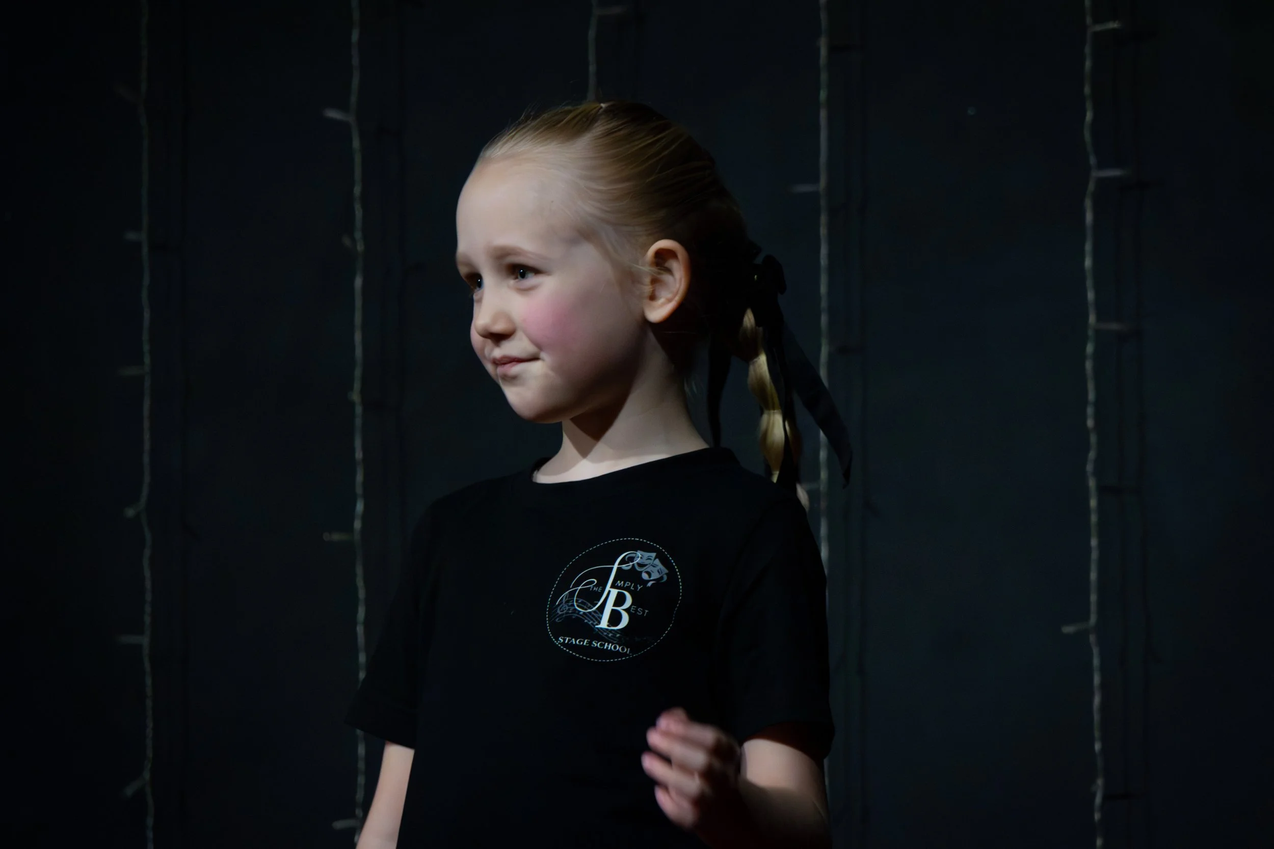 A young girl with blonde hair wearing a black stage school t-shirt, standing against a dark background with string lights.