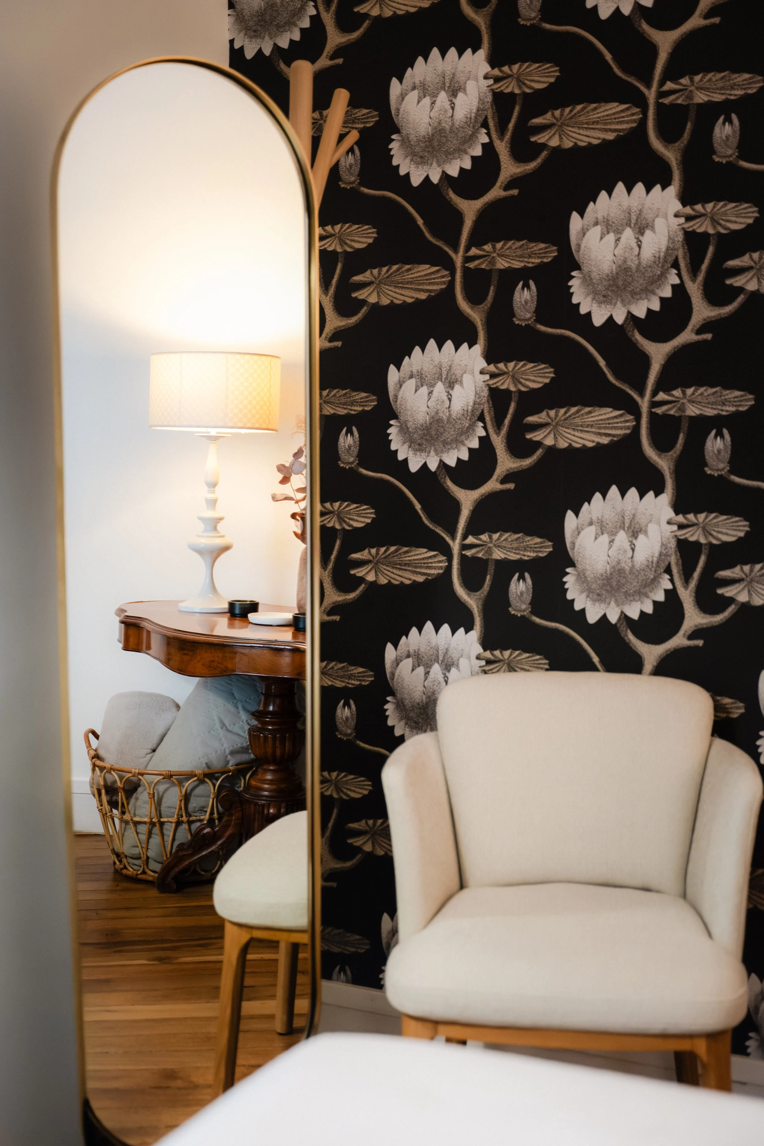 Un miroir ovale reflétant une lampe de chevet blanche avec abat-jour, une table en bois, une chaise en rotin avec coussin, et un mur à motif floral noir et blanc.