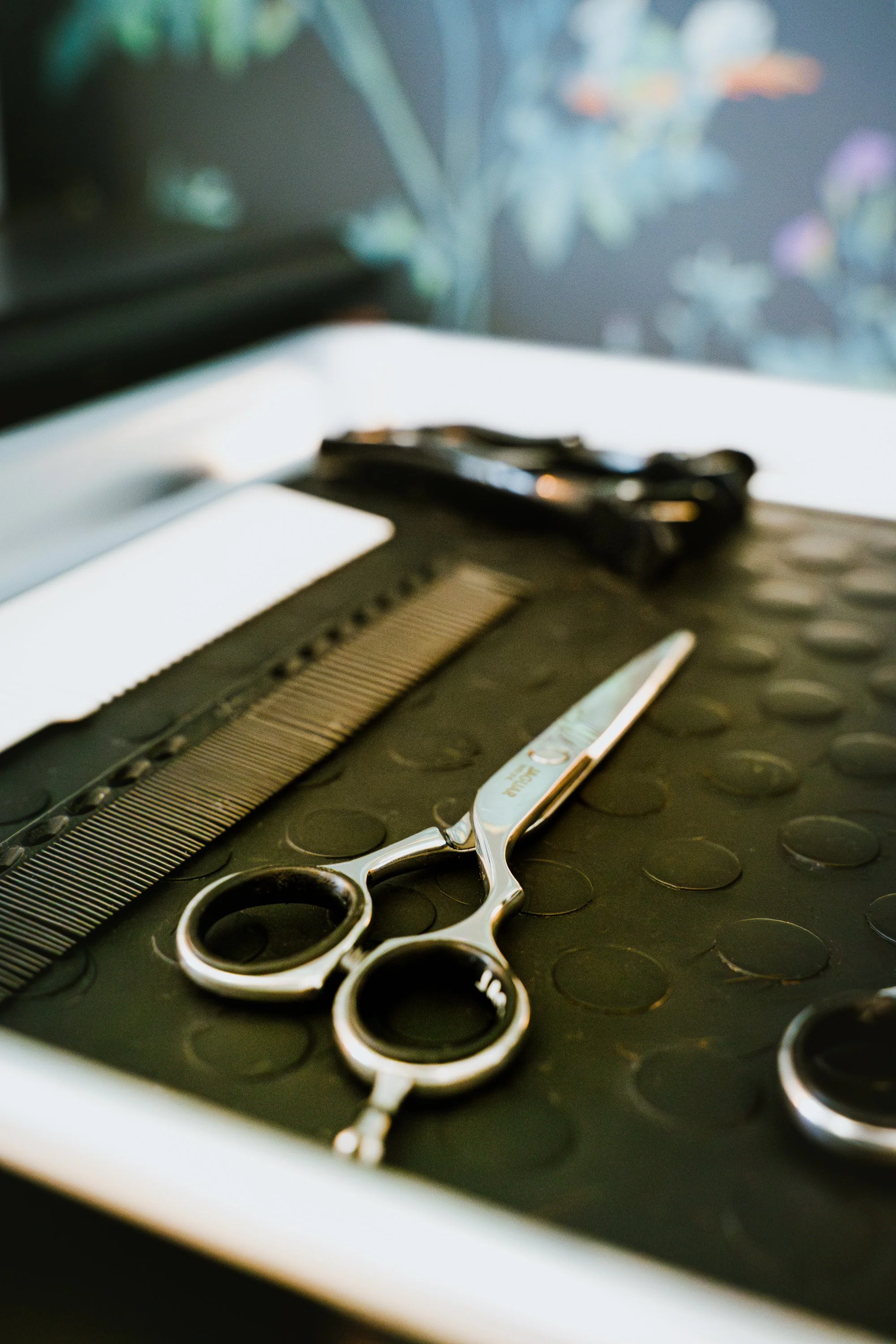Une paire de ciseaux de coiffure en inox sur une surface noire, à côté d'une tondeuse à cheveux, avec un fond flou de couleurs pastel.