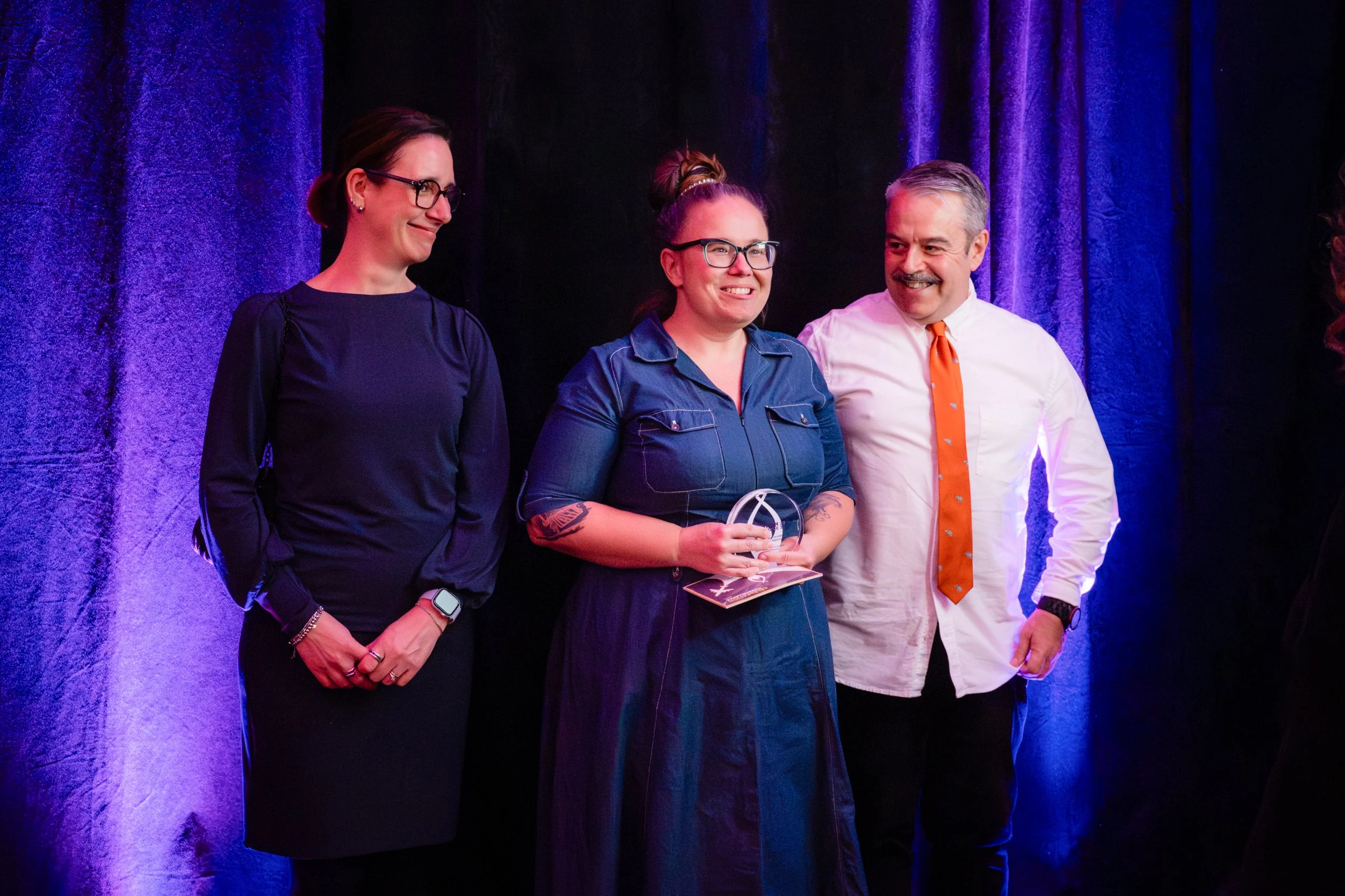 Trois personnes sont sur scène lors d'une cérémonie. La femme au centre tient un trophée et sourit, vêtue d'une chemise en denim et porte des lunettes. La femme à gauche porte une robe bleue foncée et des lunettes, la main en position de repos. L'hom
